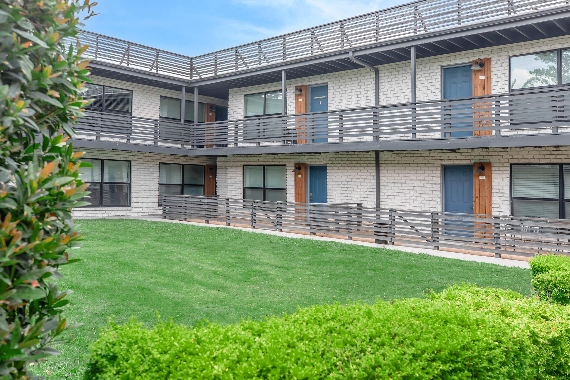 A modern apartment complex featuring multiple levels. The exterior includes a combination of white brick and dark gray accents, with balconies and doors framed by decorative railings. Lush green grass and neatly trimmed bushes are visible in the foreground, creating a welcoming outdoor space.