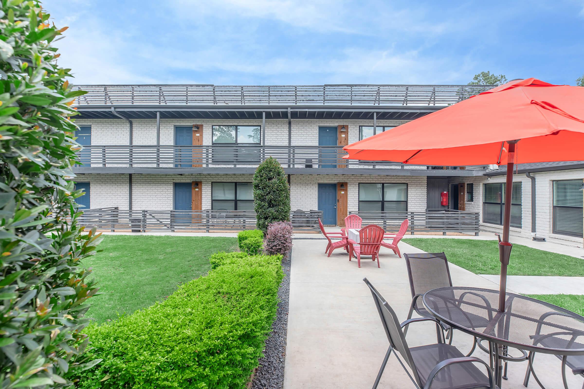 A modern hotel exterior featuring two levels of rooms with a balcony, surrounded by manicured greenery. In the foreground, a patio area includes a round table with black chairs and red Adirondack chairs under a vibrant red umbrella, inviting guests to relax outdoors.