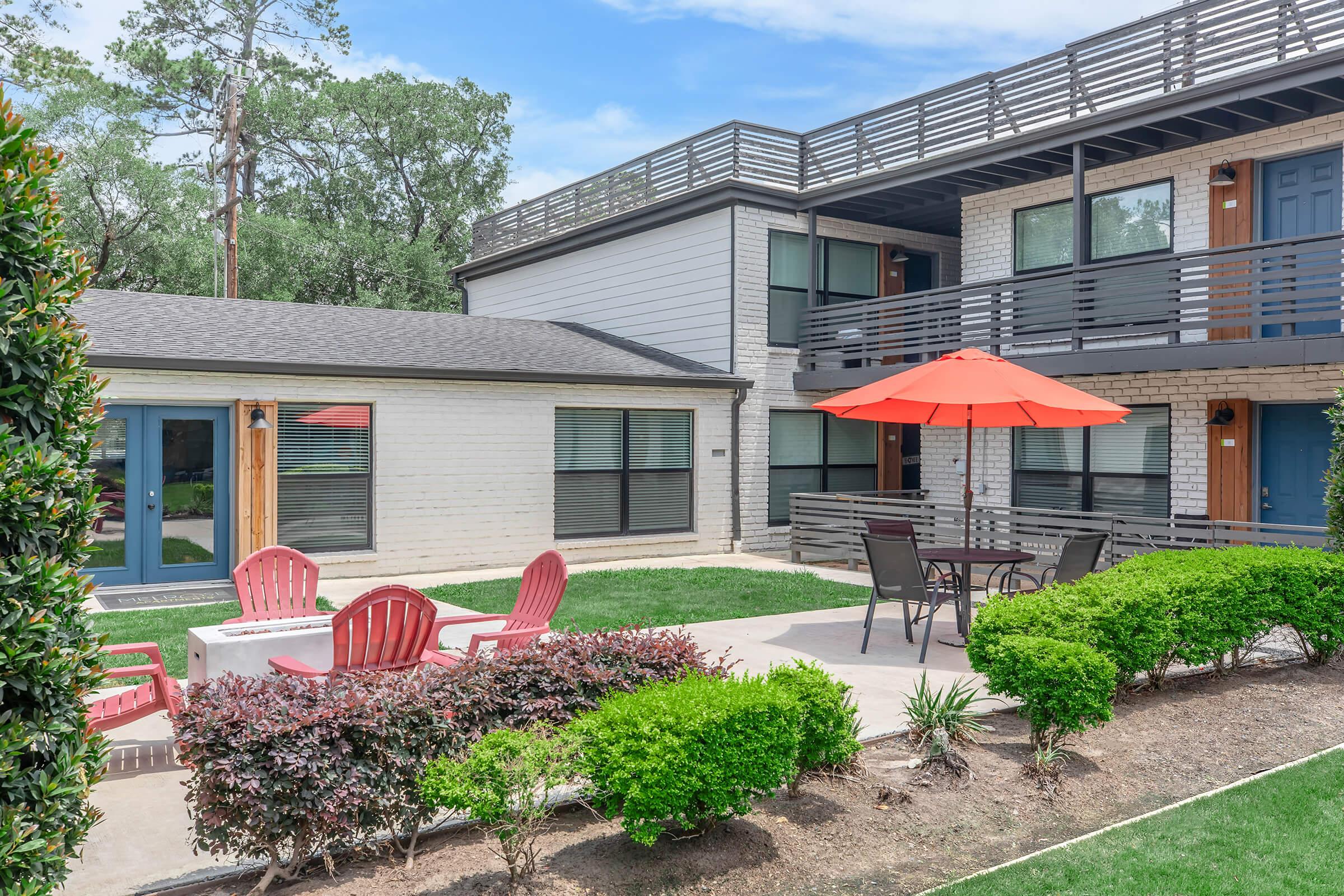 A modern multi-unit building with a landscaped area featuring green grass, bushes, and red patio furniture. There is a round table with chairs under a red umbrella and a fire pit with red chairs nearby, set against a backdrop of trees and blue sky.