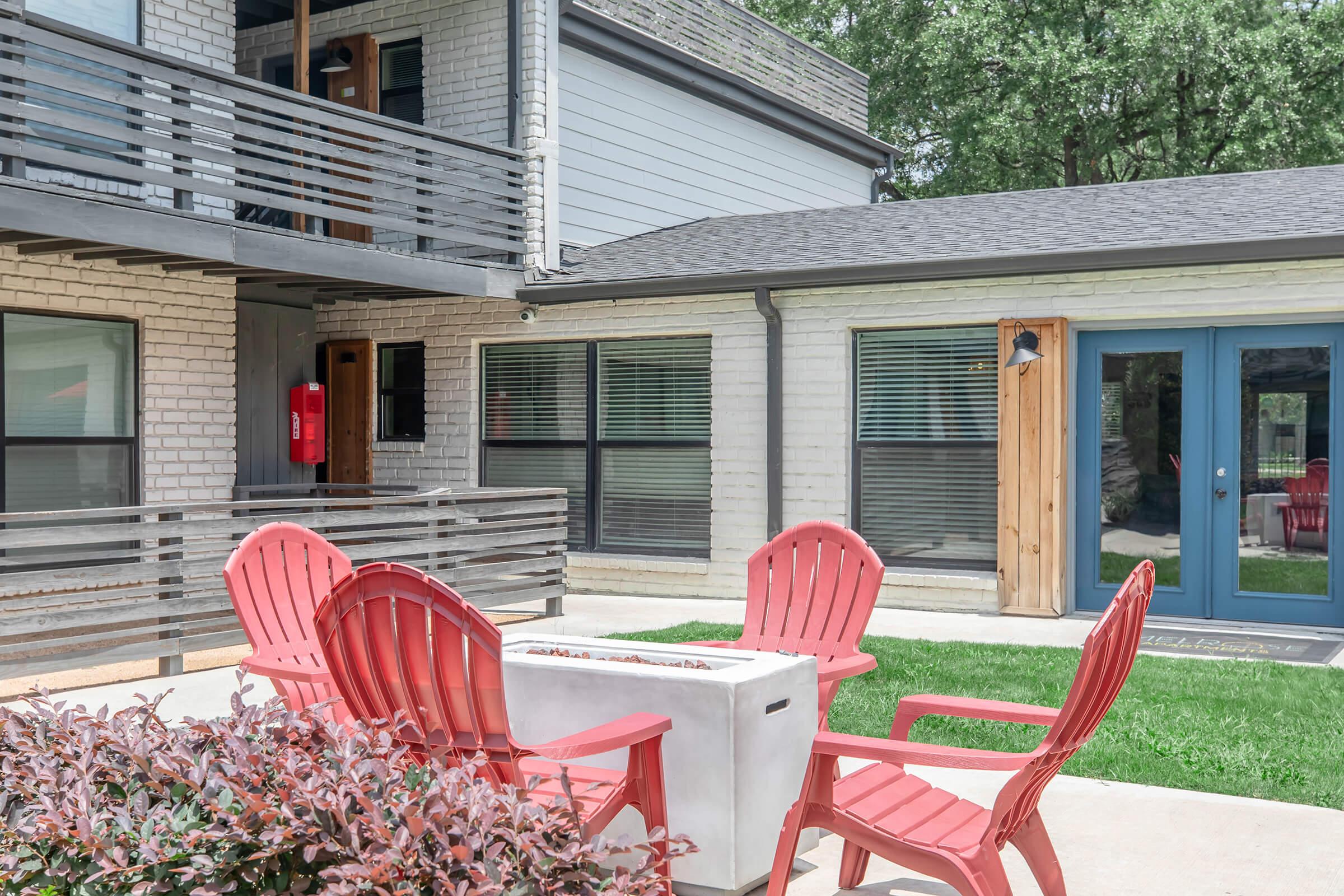 A cozy outdoor area featuring a set of red Adirondack chairs arranged around a fire pit, surrounded by green grass and a landscaped border. In the background, there are two buildings with gray exteriors, large windows, and a small wooden door. The setting is bright and inviting, perfect for relaxation.