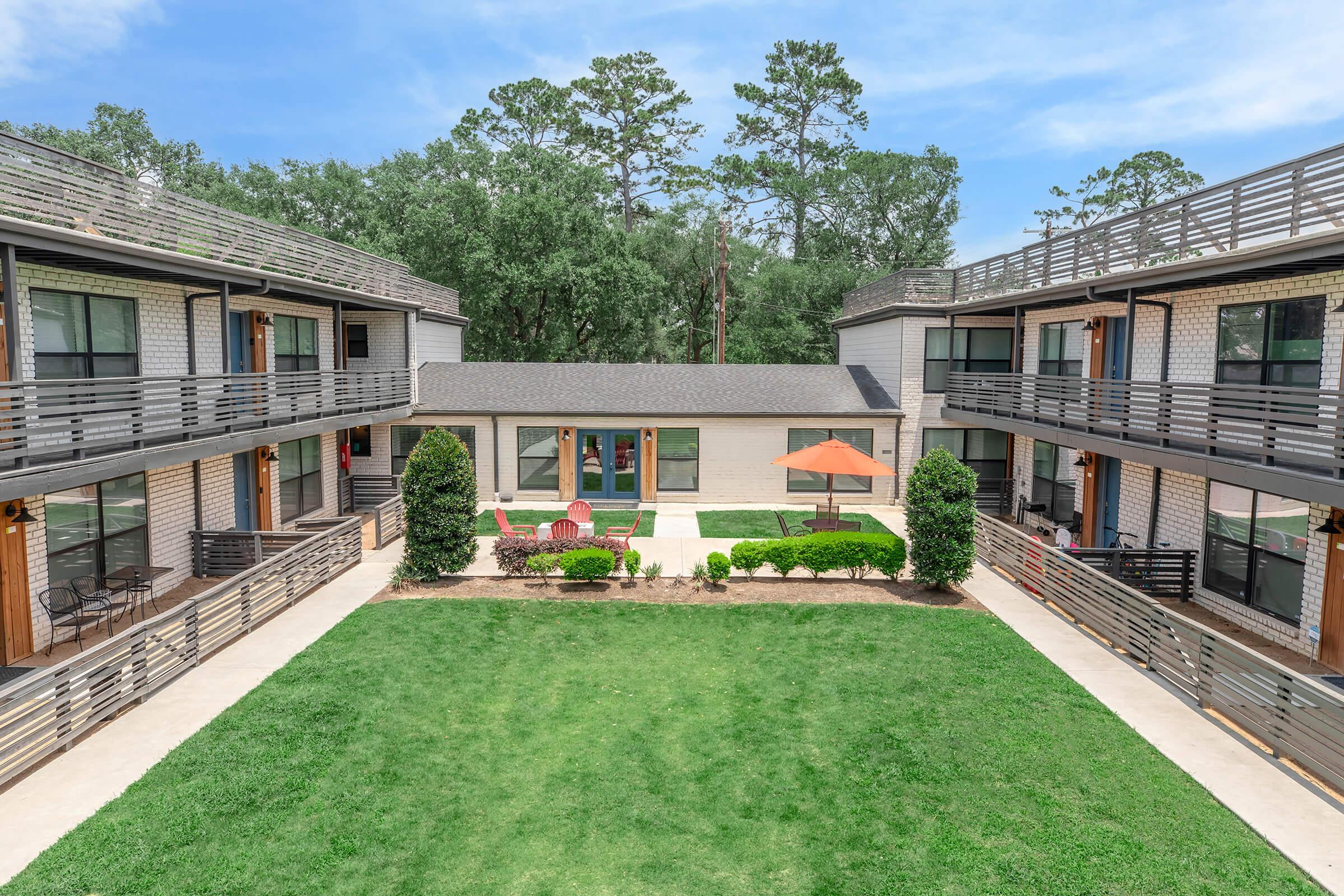 A view of a two-story apartment complex with a central green lawn and outdoor seating area. The building features multiple entrances with balconies, surrounded by trees and sunny skies. An orange umbrella provides shade over the seating area, creating a welcoming outdoor space.