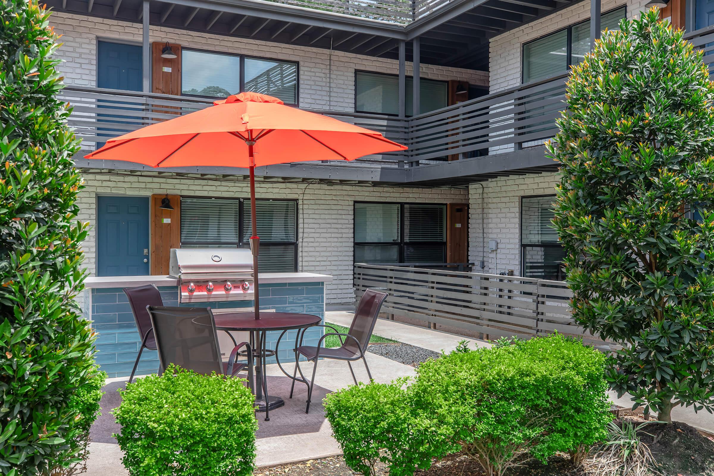 A poolside area featuring a round table with two chairs under a bright orange umbrella. There is a barbecue grill nearby. The setting includes well-maintained greenery and the exterior of a multi-story building with balconies and doors visible in the background.