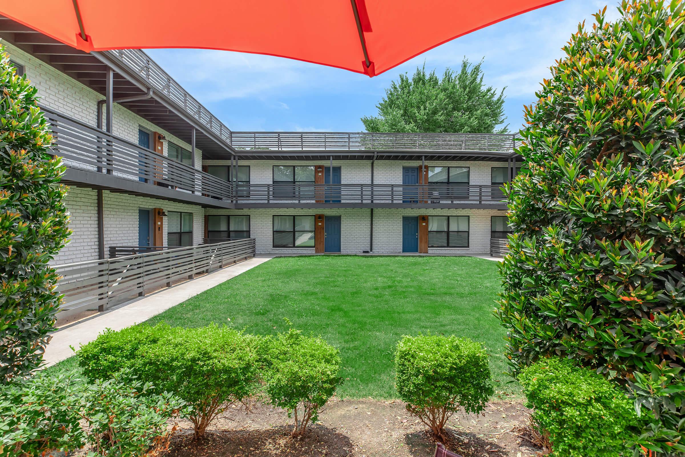 View of a courtyard between two buildings in an apartment complex, featuring green grass and neatly trimmed hedges. An orange umbrella is in the foreground, providing shade, while the buildings exhibit a modern architectural style with balconies and blue doors. Trees are visible in the background against a clear sky.