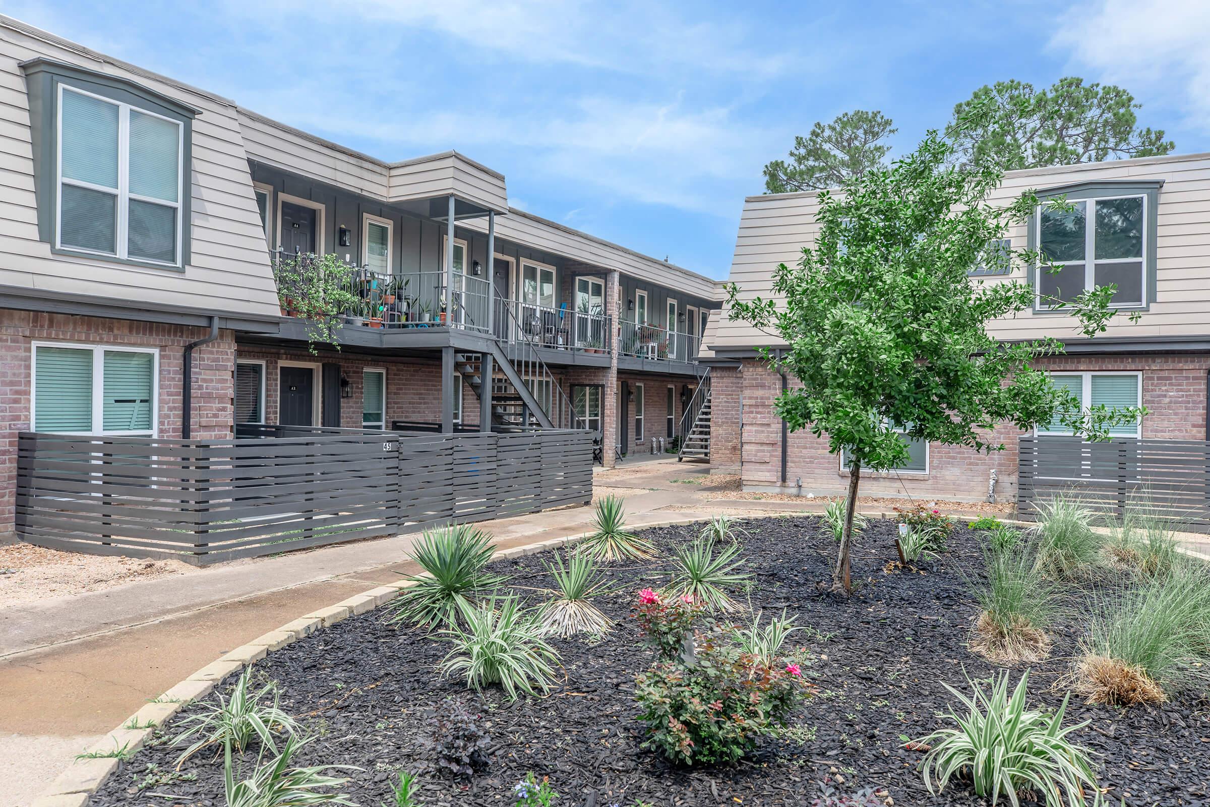 A modern apartment complex with two-story buildings surrounding a landscaped courtyard. The courtyard features low-maintenance plants, shrubs, and a tree, while the building exteriors have a mix of brick and siding. Walkways connect the entrances, and balconies are visible on the upper floor.
