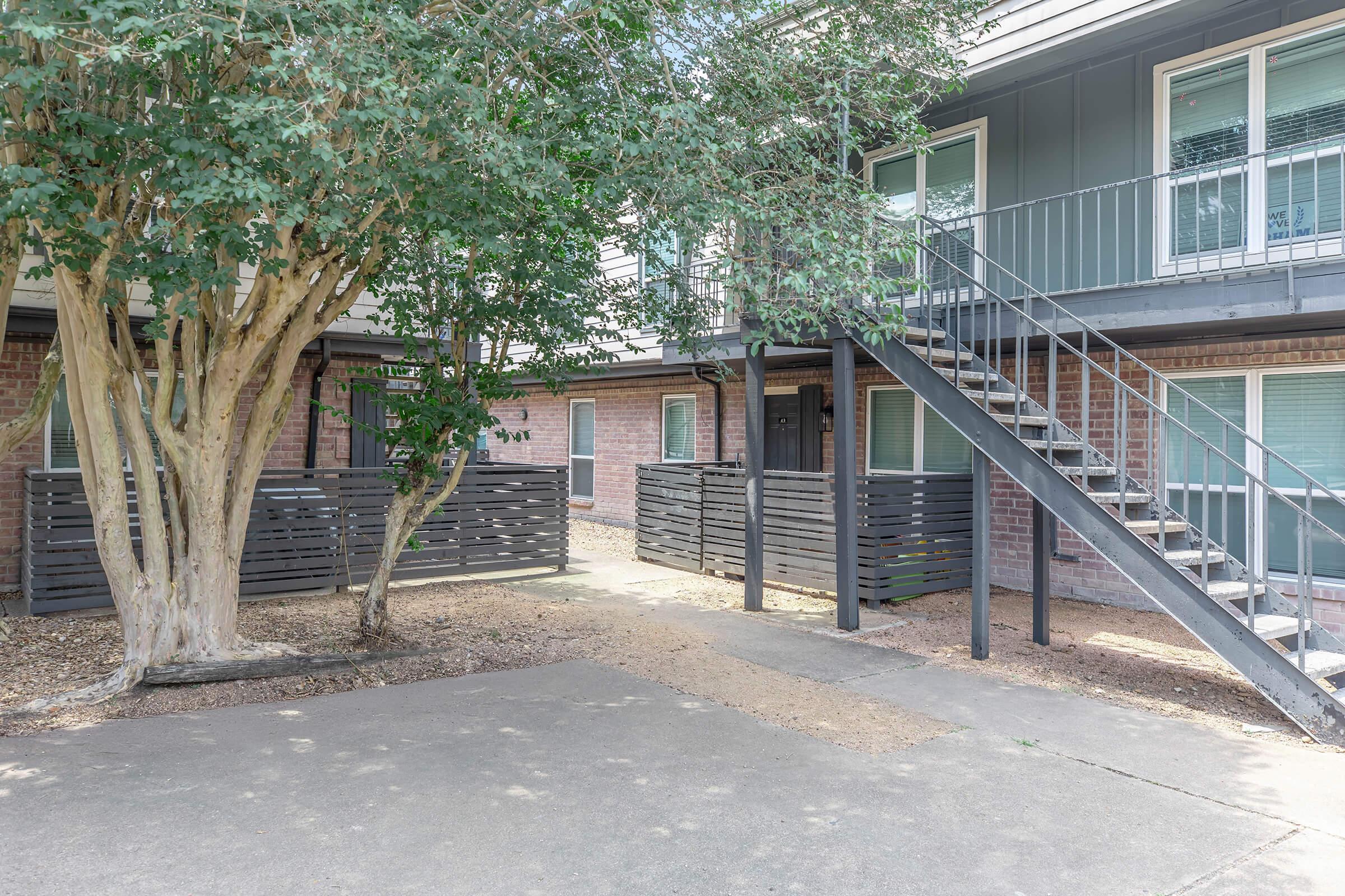 A view of a multi-unit residential building featuring a staircase leading to an upper level. Surrounding the building are small trees and wooden fences, creating a cozy courtyard atmosphere. The ground is paved, and there are windows visible on the lower level.