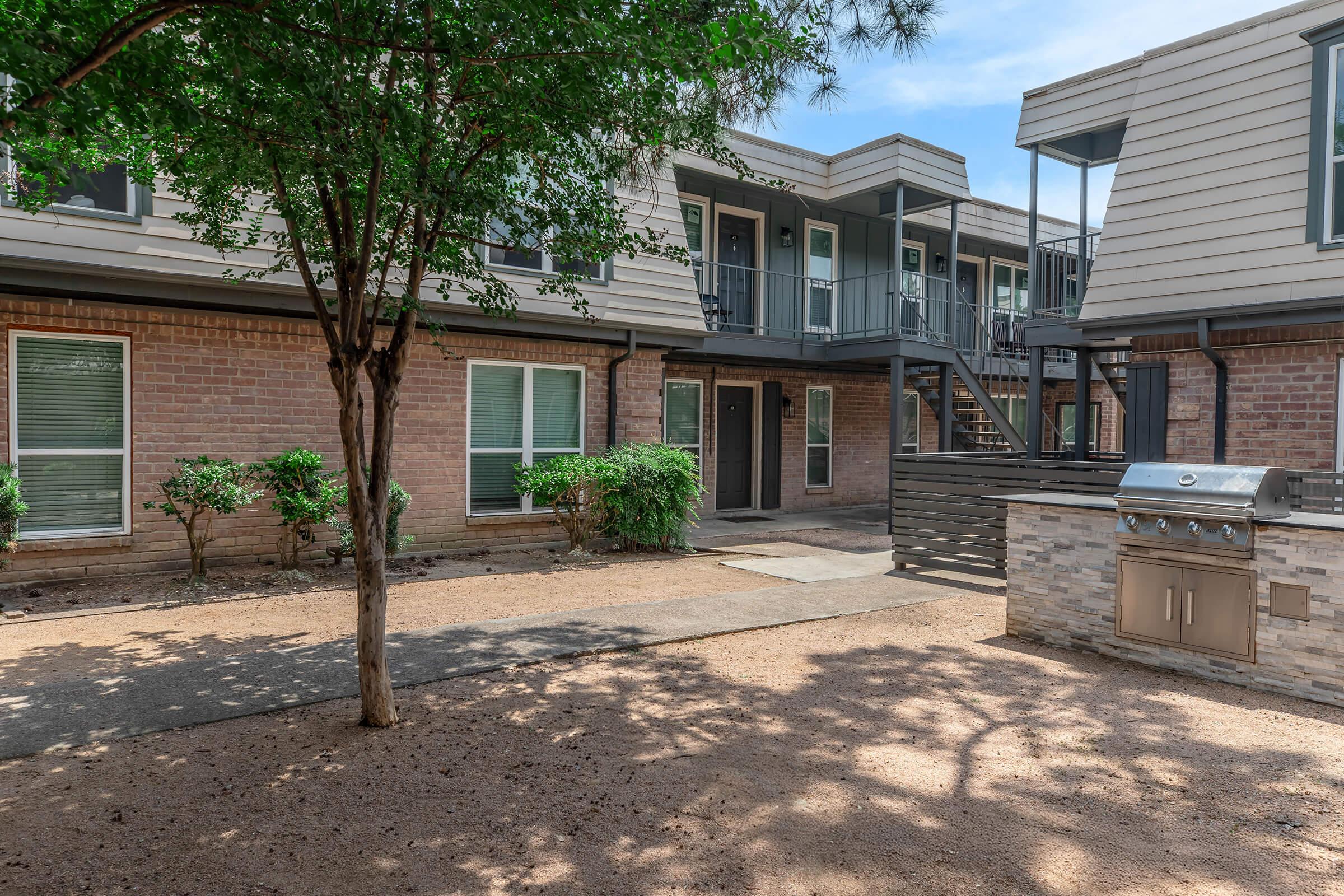 A view of a courtyard area in an apartment complex, featuring a brick building with multiple entrances, a staircase, and a grill. Lush greenery is visible, alongside a gravel path under a clear blue sky. Shadows from nearby trees create a pleasant atmosphere.