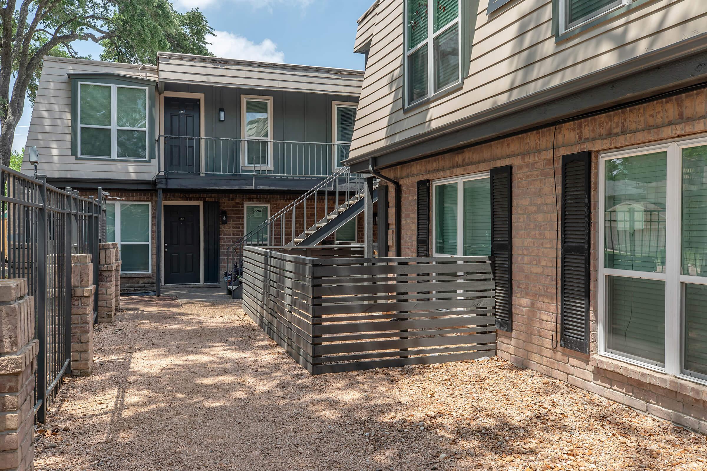 Exterior view of a multi-unit residential building featuring a deck with a wooden railing, stairs leading to an upper level, and a gravel pathway. The building has various windows, a mix of brick and siding, and is surrounded by trees, creating a pleasant living environment.