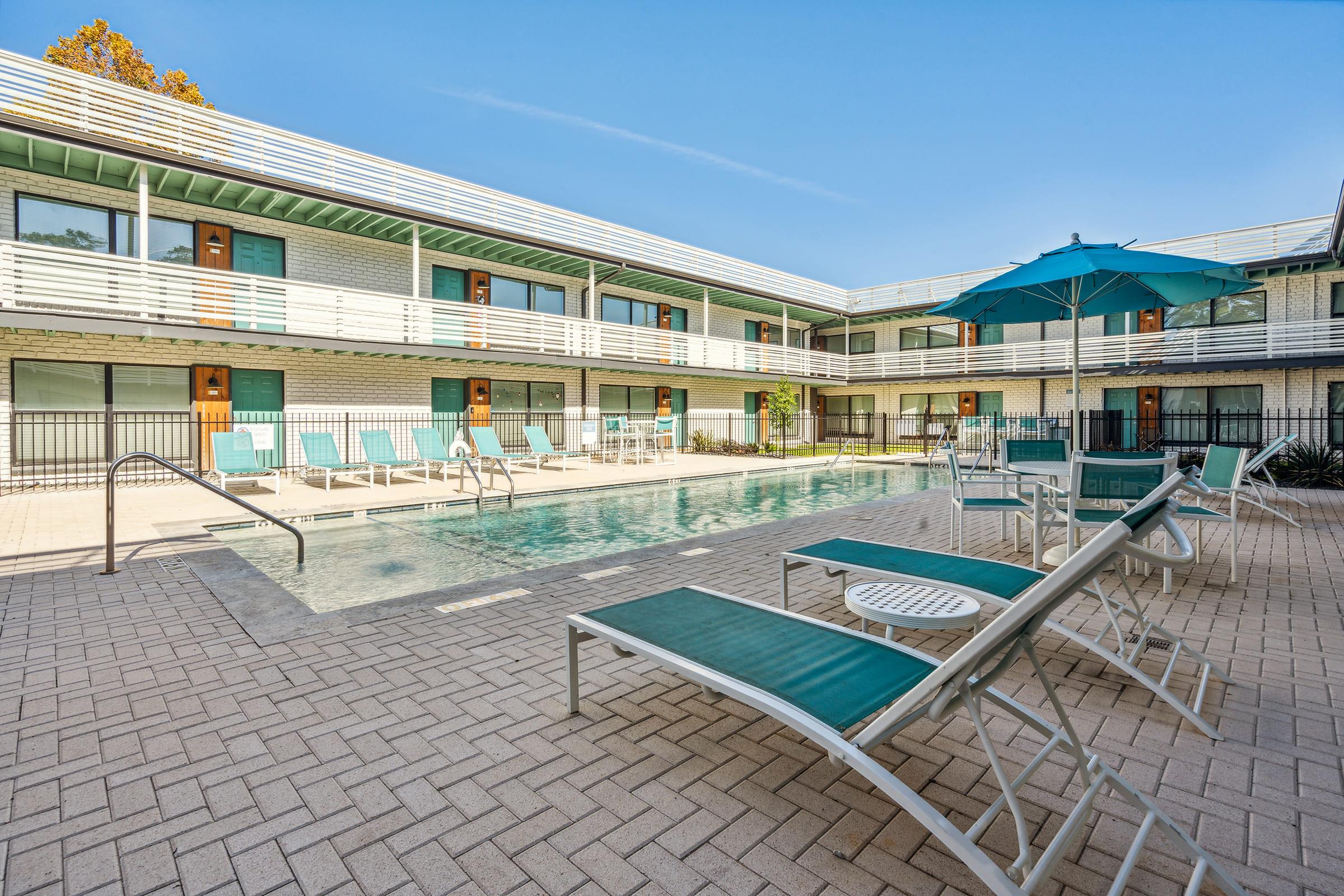 A modern hotel courtyard featuring a clear swimming pool surrounded by lounge chairs and umbrellas. The two-story building has multiple rooms accessible from the pool area, with a bright blue sky overhead and well-maintained landscaping.