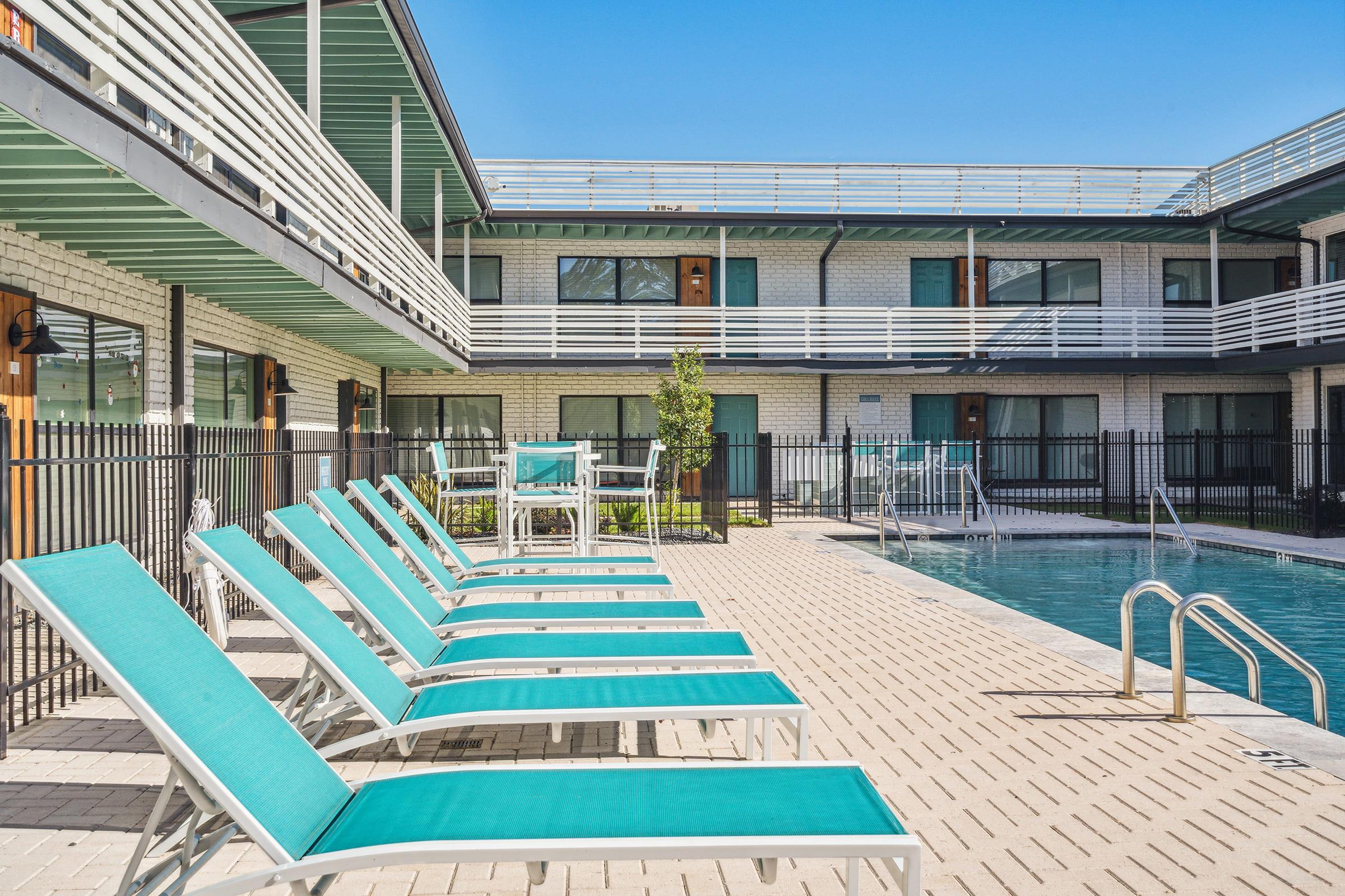 A sunny outdoor area with a swimming pool surrounded by lounge chairs. The poolside features turquoise-colored lounge chairs arranged in a line, and there are tables and chairs in the background. A modern two-story building with multiple windows is visible, set against a clear blue sky.