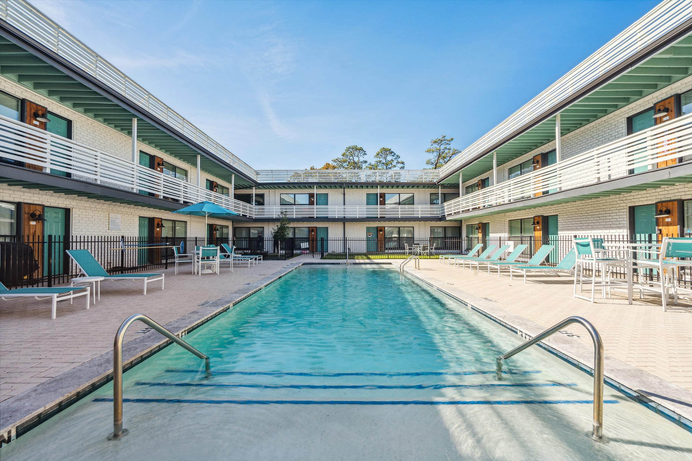 A clean and inviting motel courtyard featuring a swimming pool in the center. Surrounding the pool are lounge chairs and a few umbrellas, with two-story motel rooms visible in the background. The architecture reflects a modern, minimalist design under a clear blue sky.