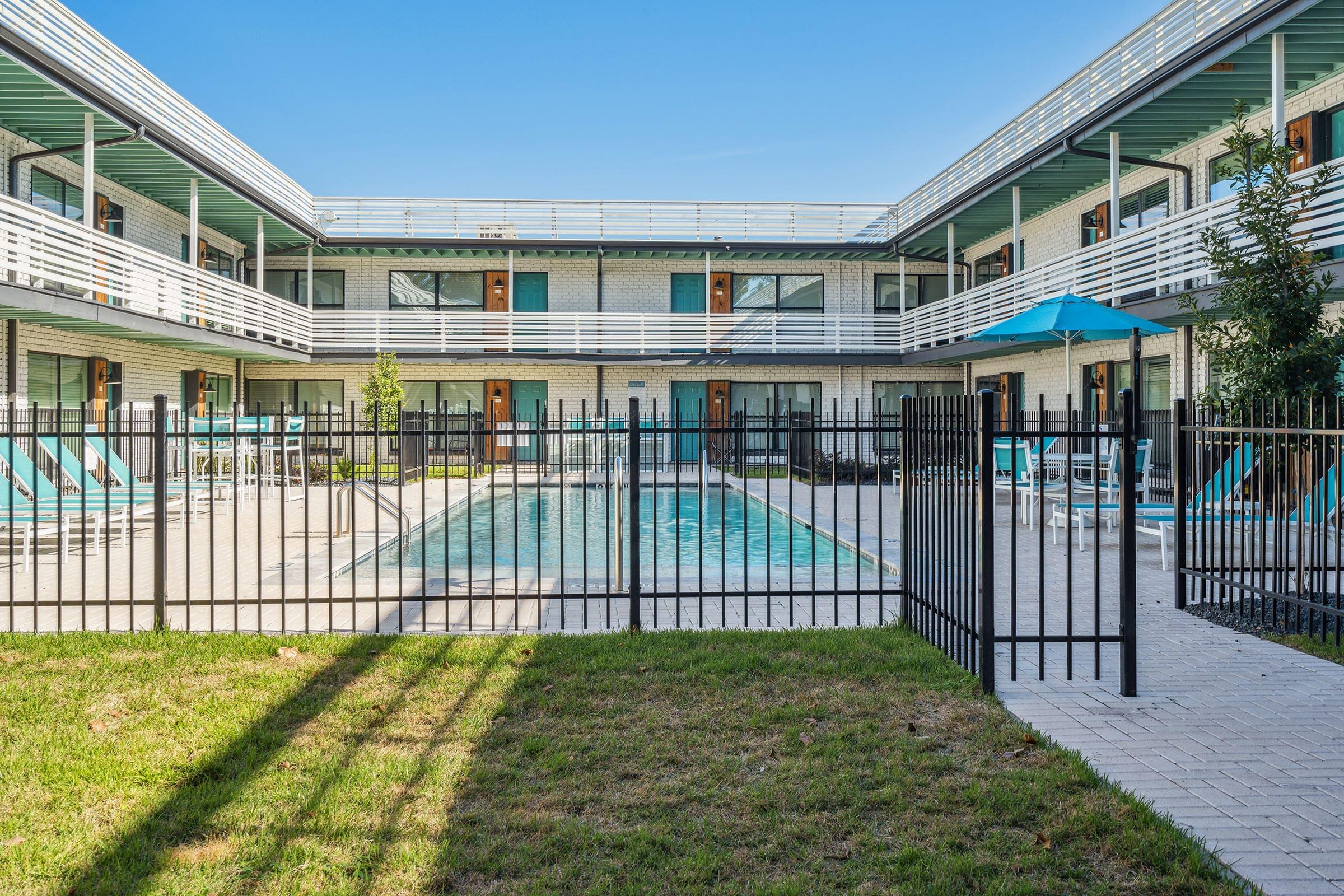 A view of a hotel courtyard featuring a swimming pool surrounded by lounge chairs. The pool is enclosed by a black metal fence, and there are multiple hotel rooms visible on the upper and lower levels, with blue umbrellas providing shade. Green grass and landscaping add to the inviting atmosphere.