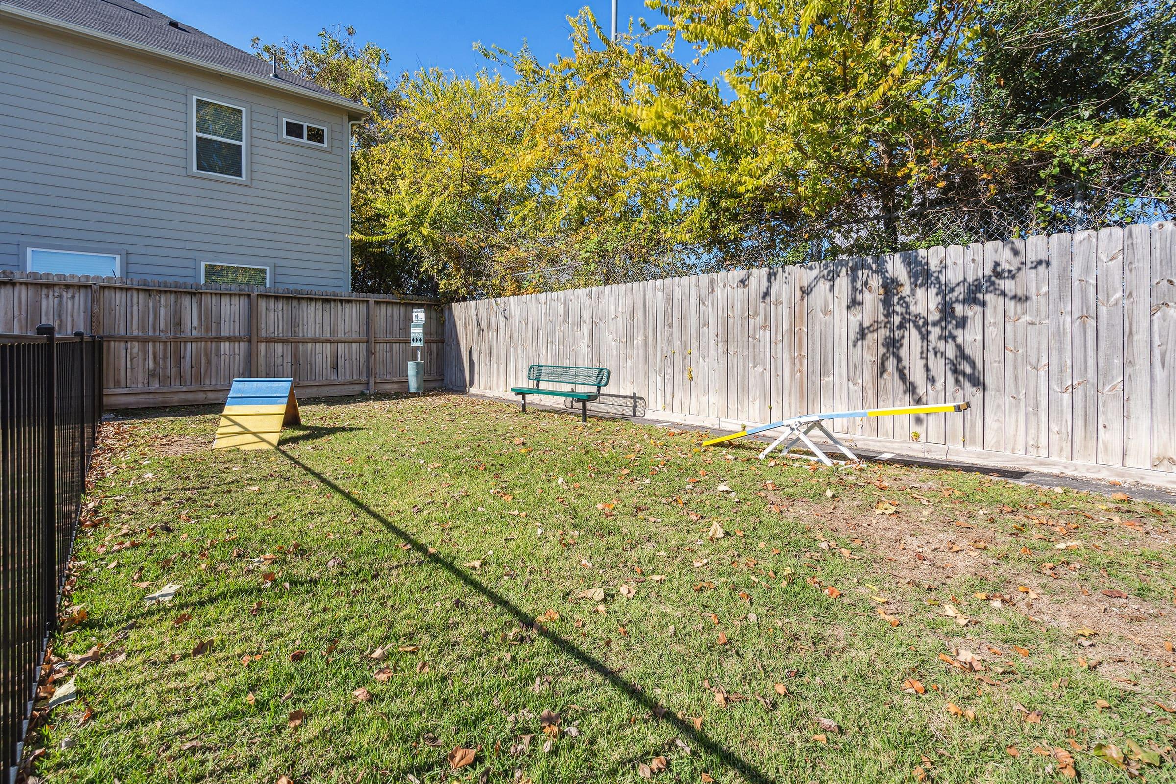 A fenced backyard featuring a grassy area with scattered leaves, a green bench, a small wooden playhouse, and a seesaw. In the background, there is a wooden fence and a house with a window visible. Bright sunlight illuminates the scene, highlighting the greenery and outdoor play equipment.