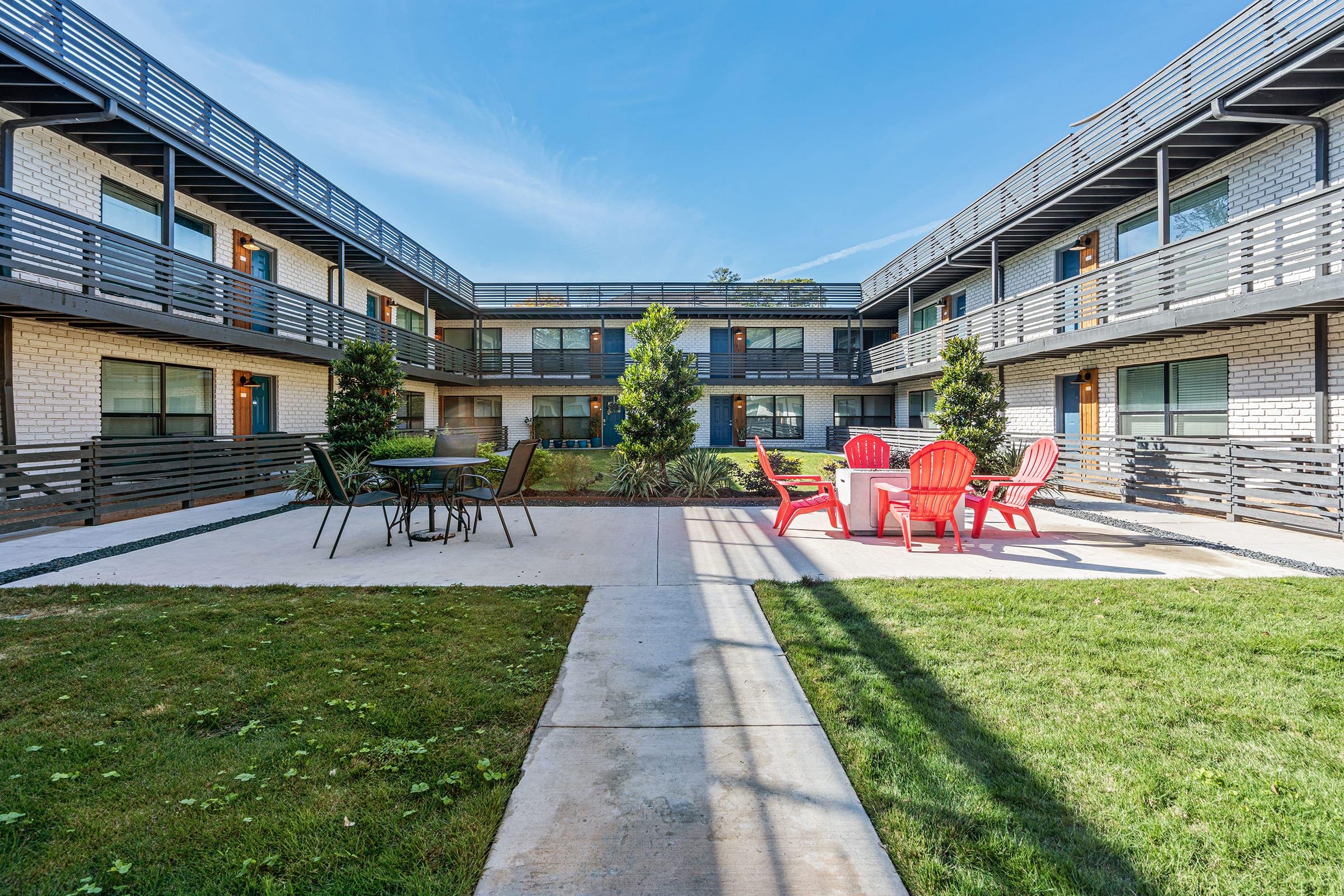 A modern courtyard with red Adirondack chairs and a table, surrounded by two-story buildings. Lush green grass and small trees enhance the inviting outdoor space, with balconies above. Clear blue sky overhead creates a bright and welcoming atmosphere.
