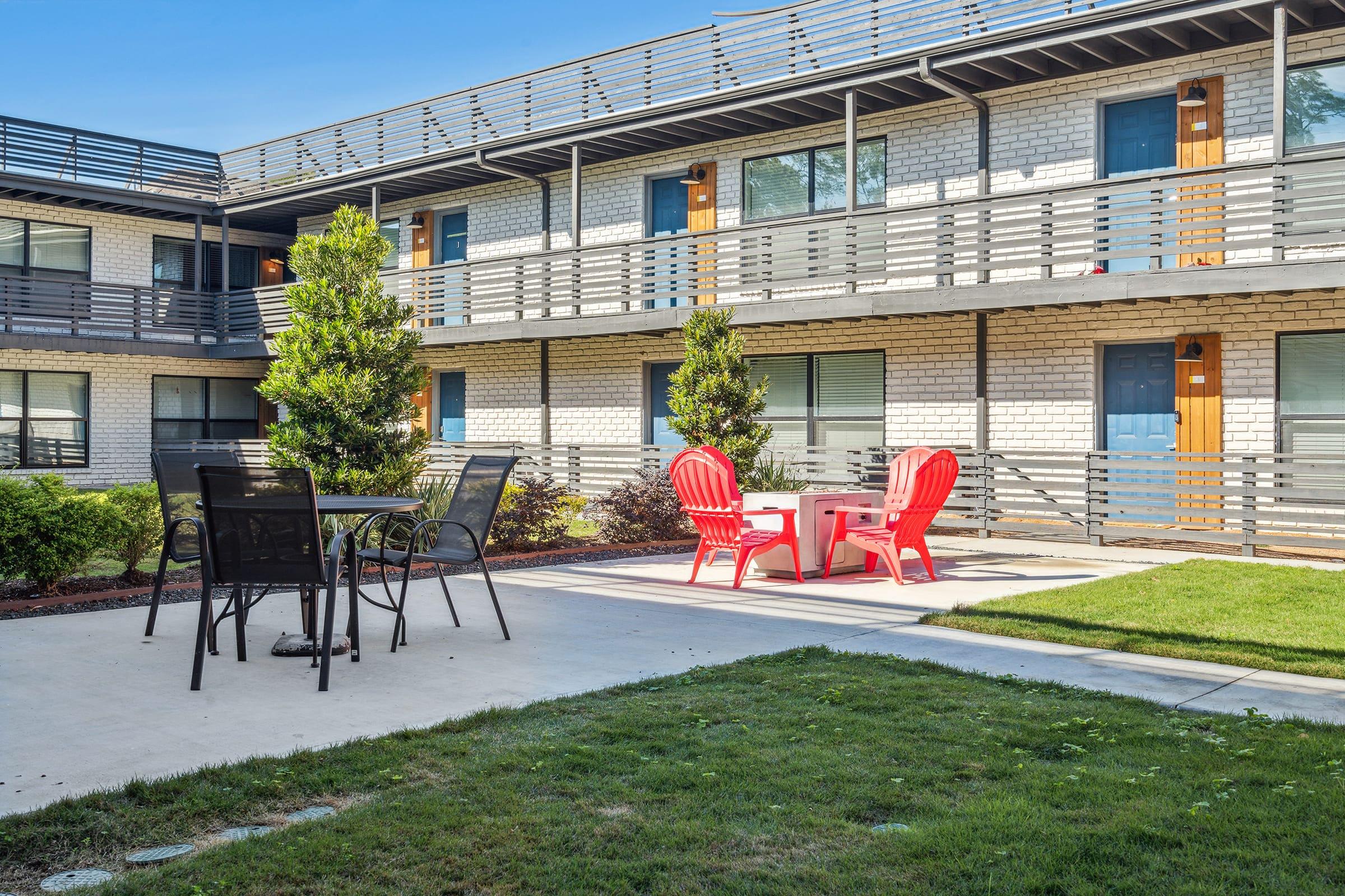 A courtyard of a modern multi-story building featuring a grassy area with a few shrubs. There are two red Adirondack chairs around a small table and a black patio set with chairs, all situated on a concrete surface. The building has multiple balconies and large windows.