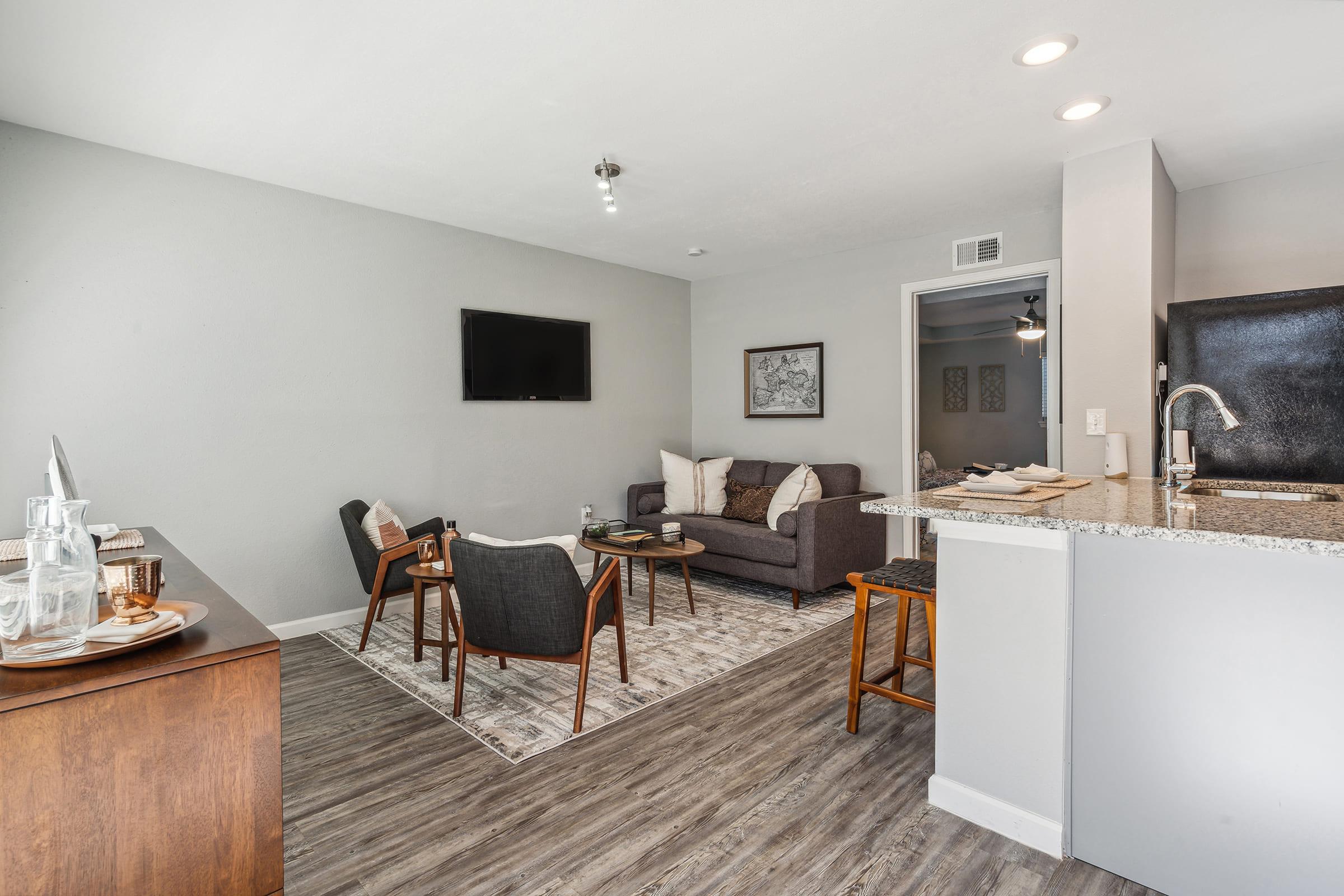 A modern living room featuring a dark gray sofa, two stylish chairs, and a wooden coffee table. A flat-screen TV is mounted on the wall. The space has light gray walls and wood-look flooring, with a small dining area visible in the background. A decorative rug adds warmth to the room.