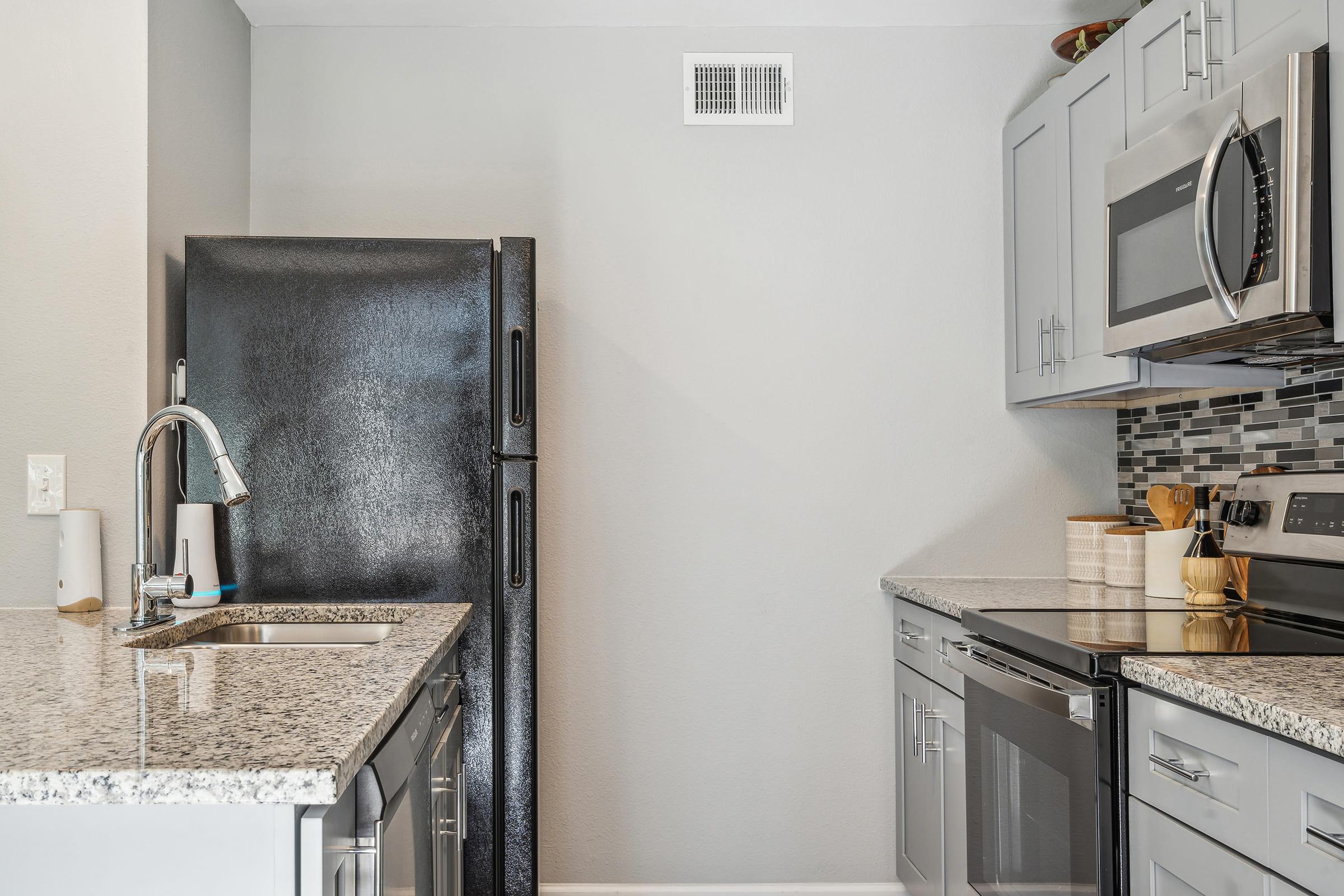 Modern kitchen featuring gray cabinetry, a black refrigerator, and stainless steel appliances. The countertop is made of speckled granite, complemented by a sleek sink and a neutral wall. Decorative items on the countertop add a touch of warmth to the contemporary design.