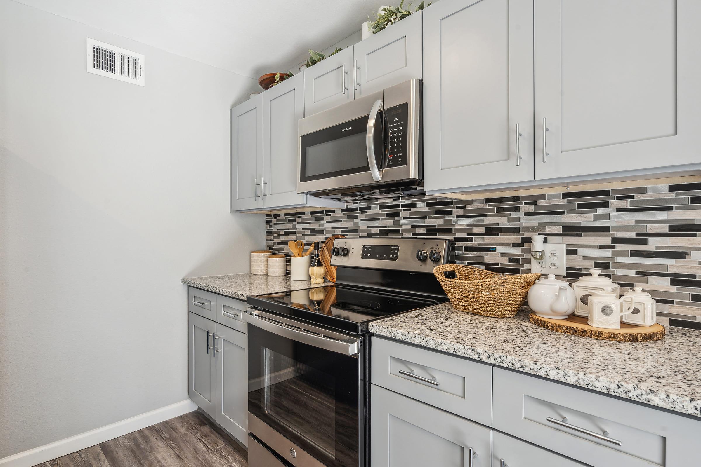 Modern kitchen featuring gray cabinets, a stainless steel microwave, an electric stove, and a microwave. The backsplash is a mix of gray and black tiles, and the countertop is light-colored granite. A woven basket and white dishware are neatly arranged on the countertop.