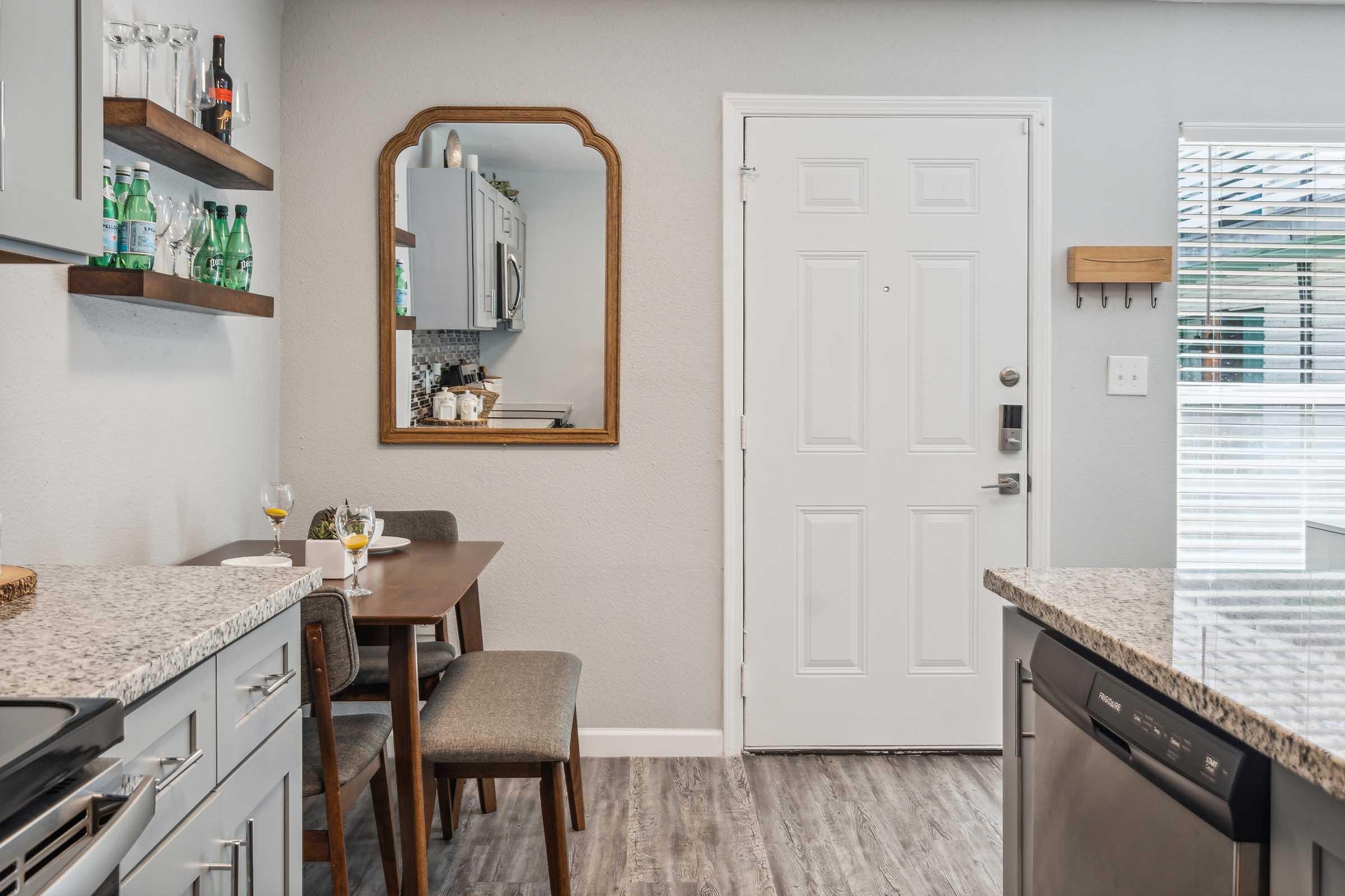 A modern kitchen area featuring a gray color scheme. There is a small dining table with two chairs, a mirrored wall hanging, and shelves displaying glassware. The doorway is visible, leading outside, with a light-colored door and a welcome sign. A countertop with a sink and dishwasher is also present.