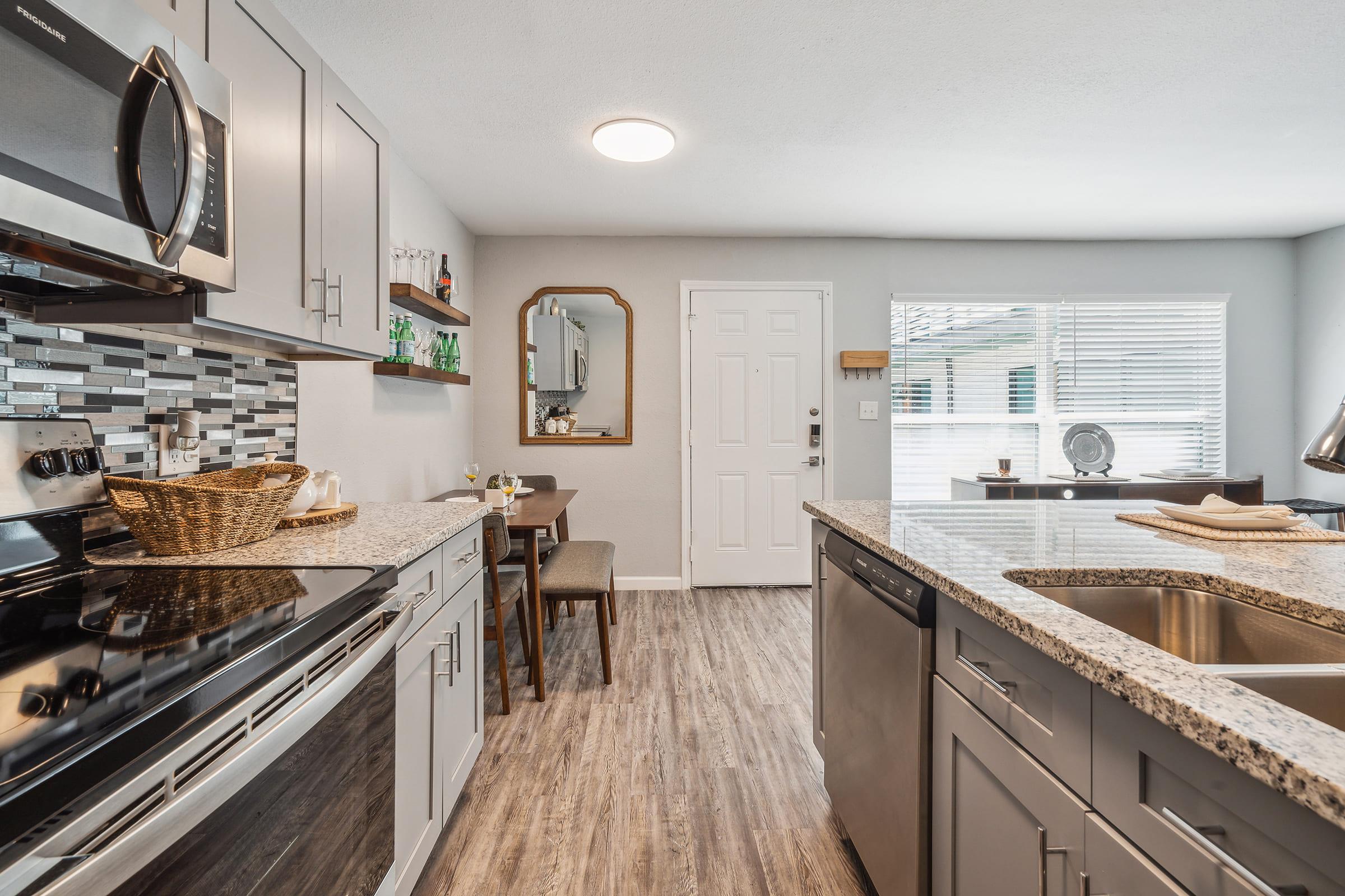 Modern kitchen featuring stainless steel appliances, gray cabinetry, and a granite countertop. A dining area with a wooden table and two chairs is visible. Natural light streams in through a window, and a mirror hangs on the wall. The flooring is a wood-like laminate, creating a warm and inviting atmosphere.