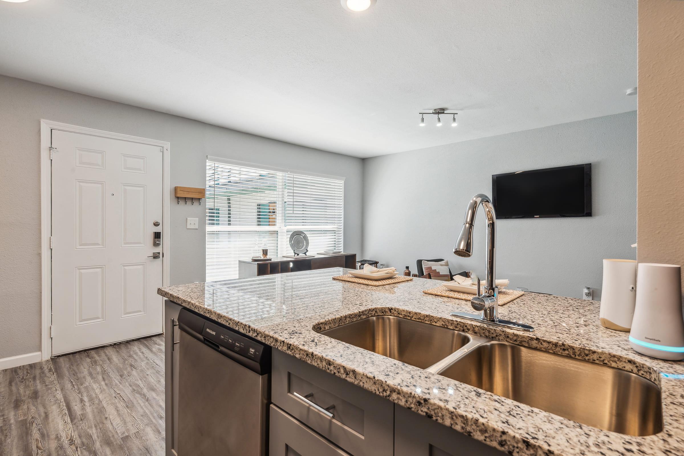 A modern kitchen with a granite countertop and double sink. It features a dishwasher and is open to a living area with a window allowing natural light. There's a small table set for dining, a wall-mounted TV, and minimalistic decor in neutral colors.