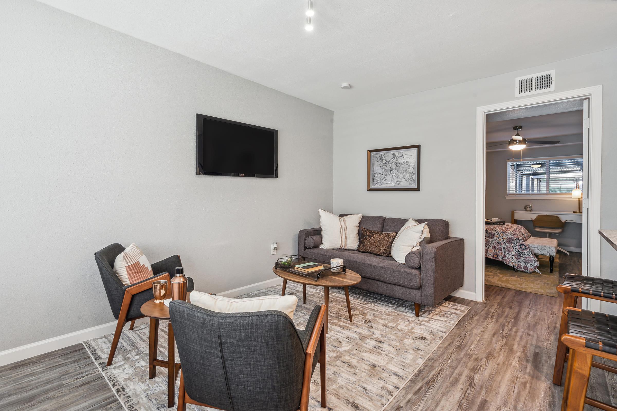A cozy living room featuring a gray sofa, two armchairs, a coffee table, and a wall-mounted TV. A patterned rug covers the floor, and a framed artwork hangs on the wall. A doorway leads to a bedroom area in the background, with soft lighting creating an inviting atmosphere.