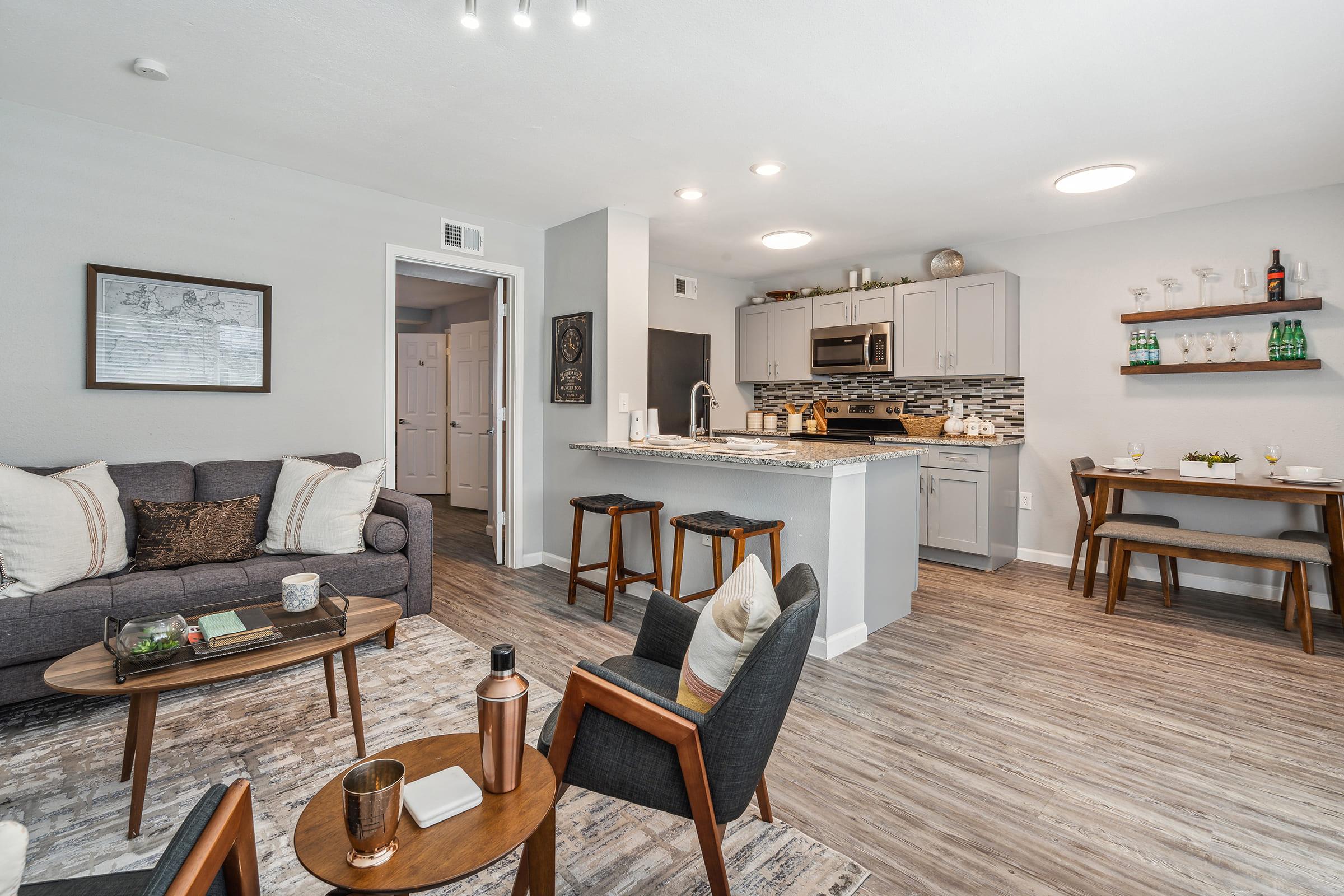 A modern, stylish living area featuring a cozy gray sofa with decorative pillows, a wooden coffee table, and a sleek kitchen with stainless steel appliances and a breakfast bar with stools. The decor includes a framed artwork on the wall, and natural light brightens the space, creating a welcoming atmosphere.