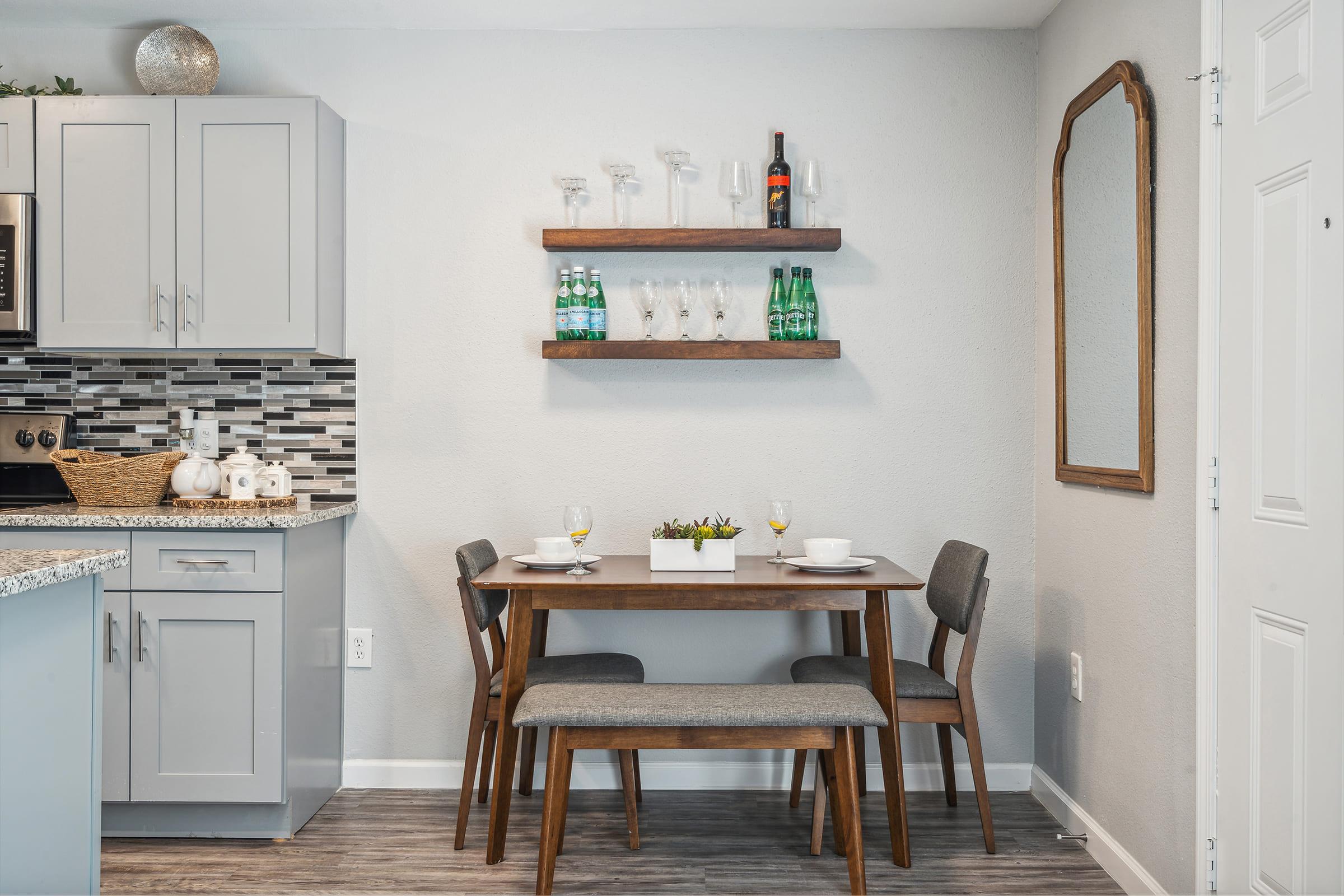 A cozy dining area featuring a wooden table with two chairs and a bench. On the table are two plates and glasses. A wooden shelf on the wall holds glasses and a bottle. A large mirror is mounted next to the door, with a light gray wall and modern cabinetry in the background, creating a warm, inviting atmosphere.