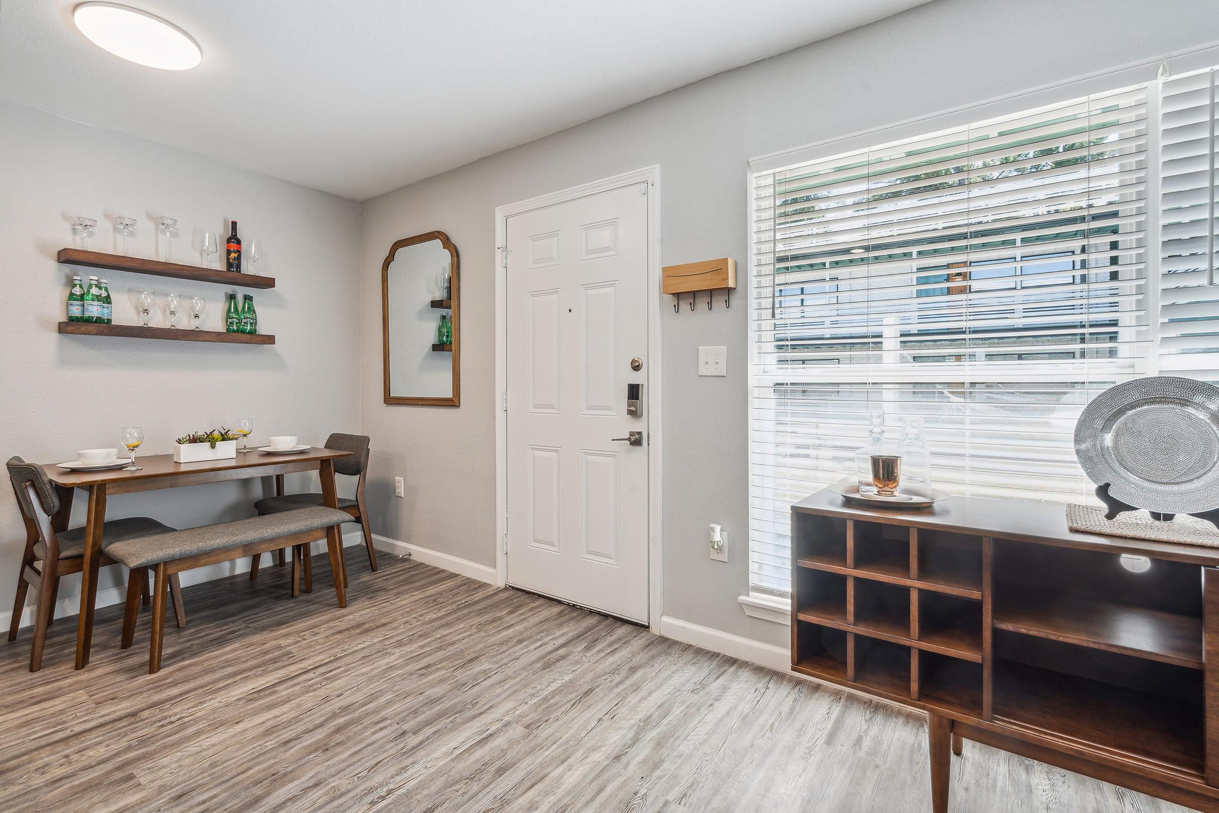A cozy dining area featuring a small table set for two, positioned near a door and a window with blinds. The wall displays a mirror and shelves holding bottles, while a sideboard with decorative items complements the warm, modern aesthetic of the space.
