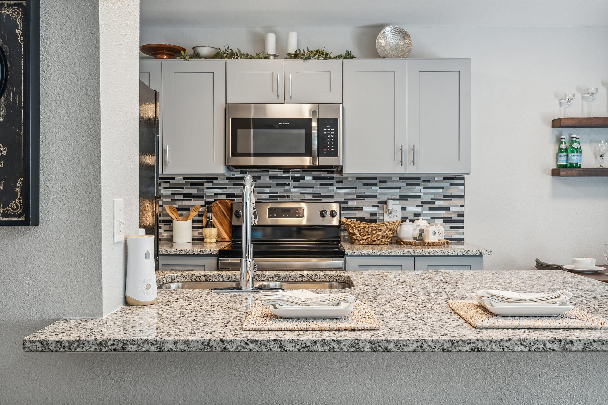 Modern kitchen with gray cabinets, stainless steel appliances, and a decorative backsplash. The countertop is made of granite, featuring plates and woven placemats. Decorative items include a fruit basket and bottles on open shelves. The space is well-lit and inviting.