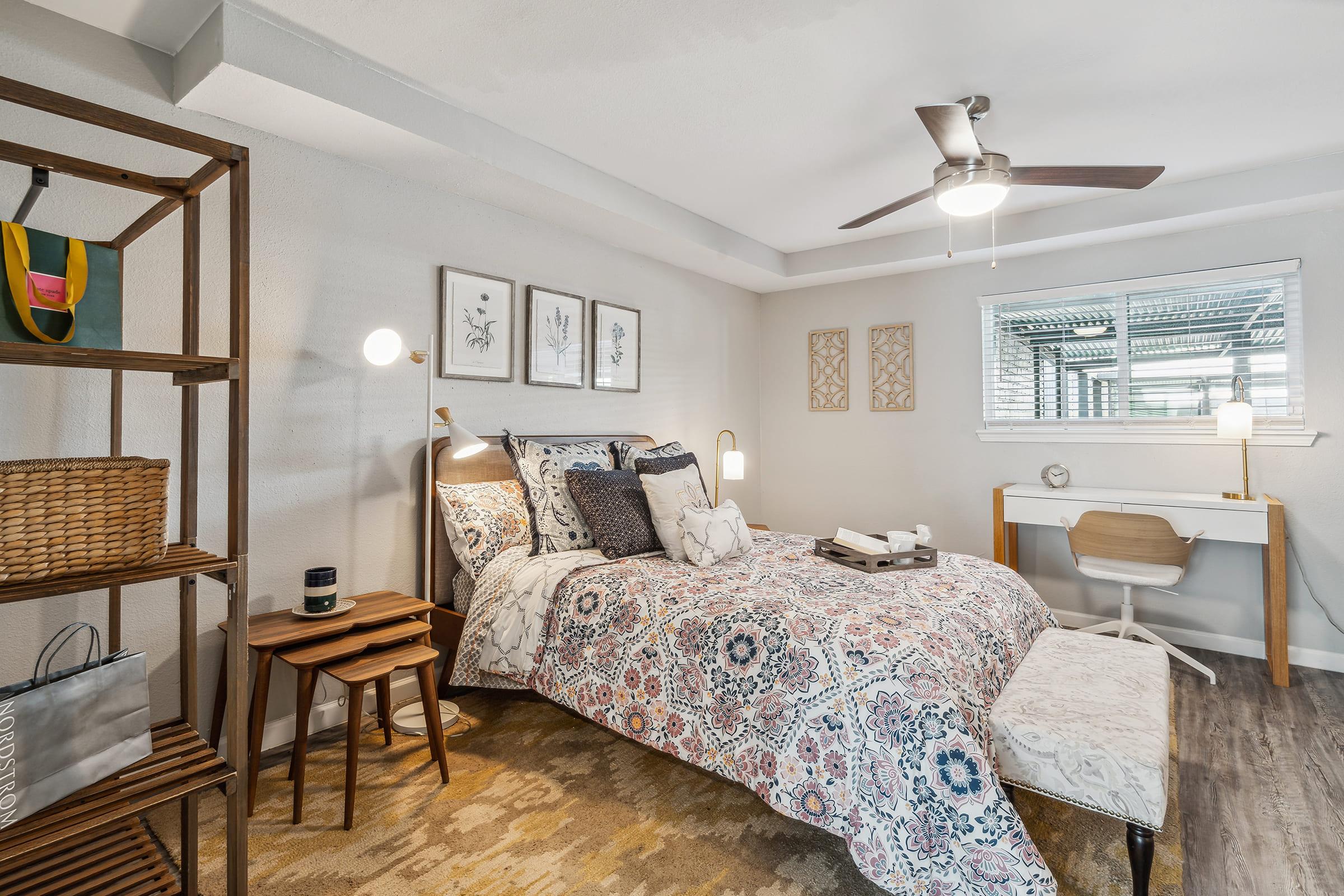 Cozy bedroom featuring a patterned bedspread, decorative pillows, and wood furniture. There is a bedside table with a lamp, a writing desk with a chair, and a window with blinds. A shelf displays a basket and a decorative bag. The room has a warm, inviting atmosphere with neutral tones and natural light.