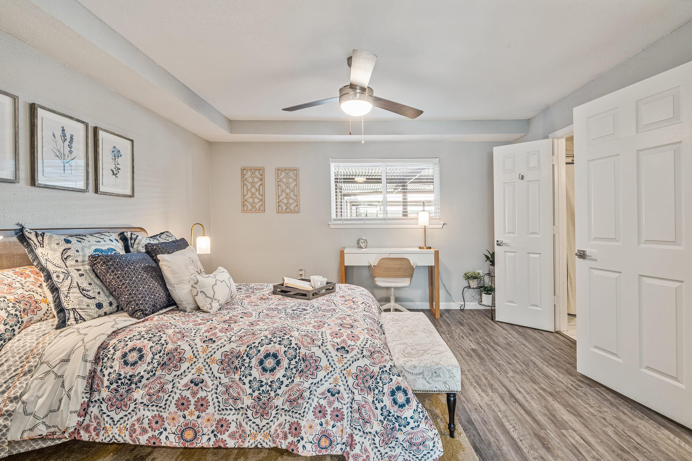 Cozy bedroom featuring a colorful patterned bedding set on a queen-sized bed, two decorative wall artworks, a bedside lamp, and a small table with a chair. A ceiling fan hangs above, and a doorway leads to another room. Natural light enters through a window, enhancing the warm ambiance.