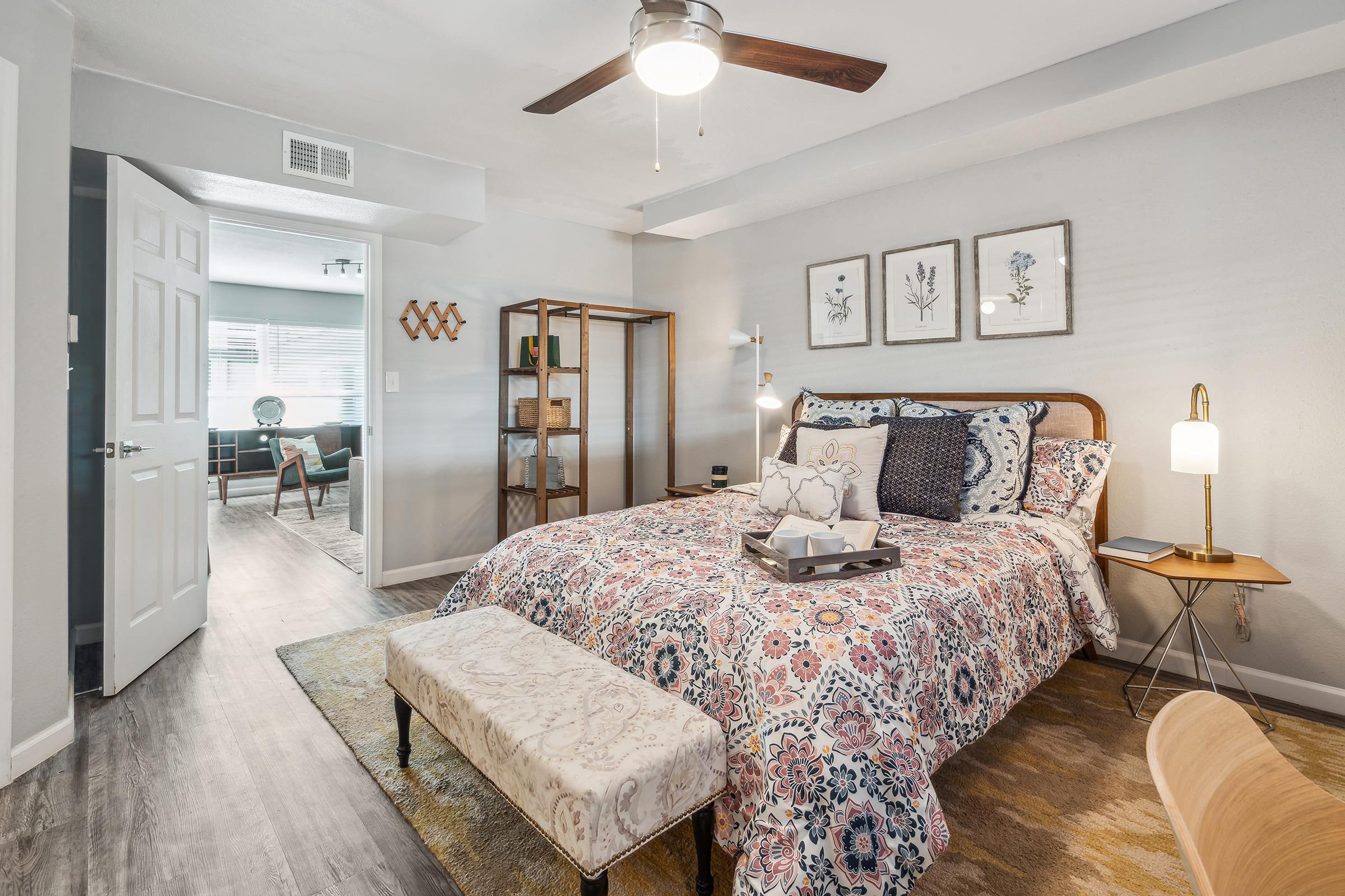 A cozy bedroom featuring a queen-sized bed with a colorful floral comforter, a decorative bench at the foot, and a stylish bedside lamp. The room has a ceiling fan and wall art consisting of framed botanical prints. A doorway leads to a living area with large windows and modern furnishings.