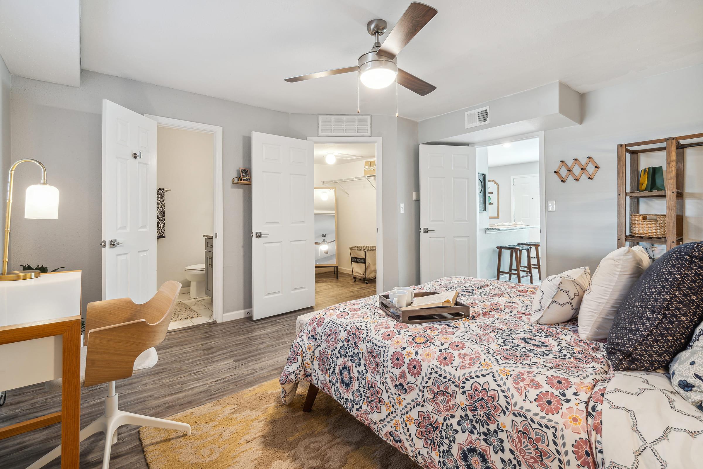 A cozy bedroom featuring a colorful floral patterned bedspread, a wooden desk with a modern chair, and a ceiling fan. Two open doors lead to a bathroom and a walk-in closet. Neutral-colored walls and a warm rug complete the inviting atmosphere of the room.
