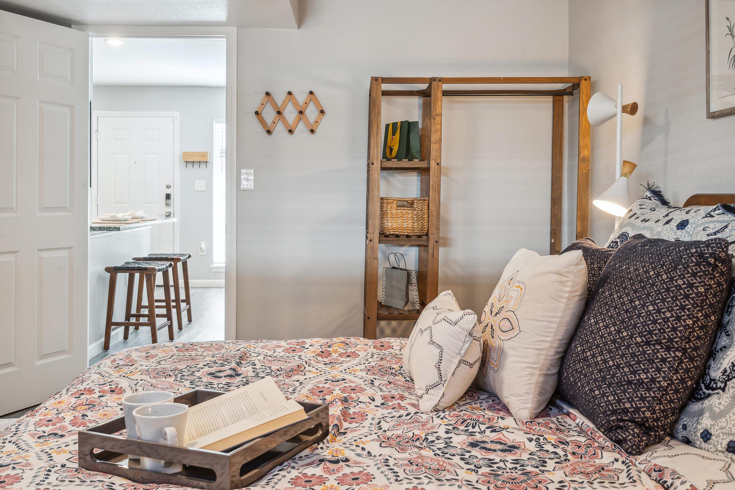 Cozy bedroom featuring a floral patterned bedspread with decorative pillows, a bedside tray with a book and mug, a wooden clothing rack with woven baskets, and a soft lamp. A doorway in the background leads to a bright, modern living area.