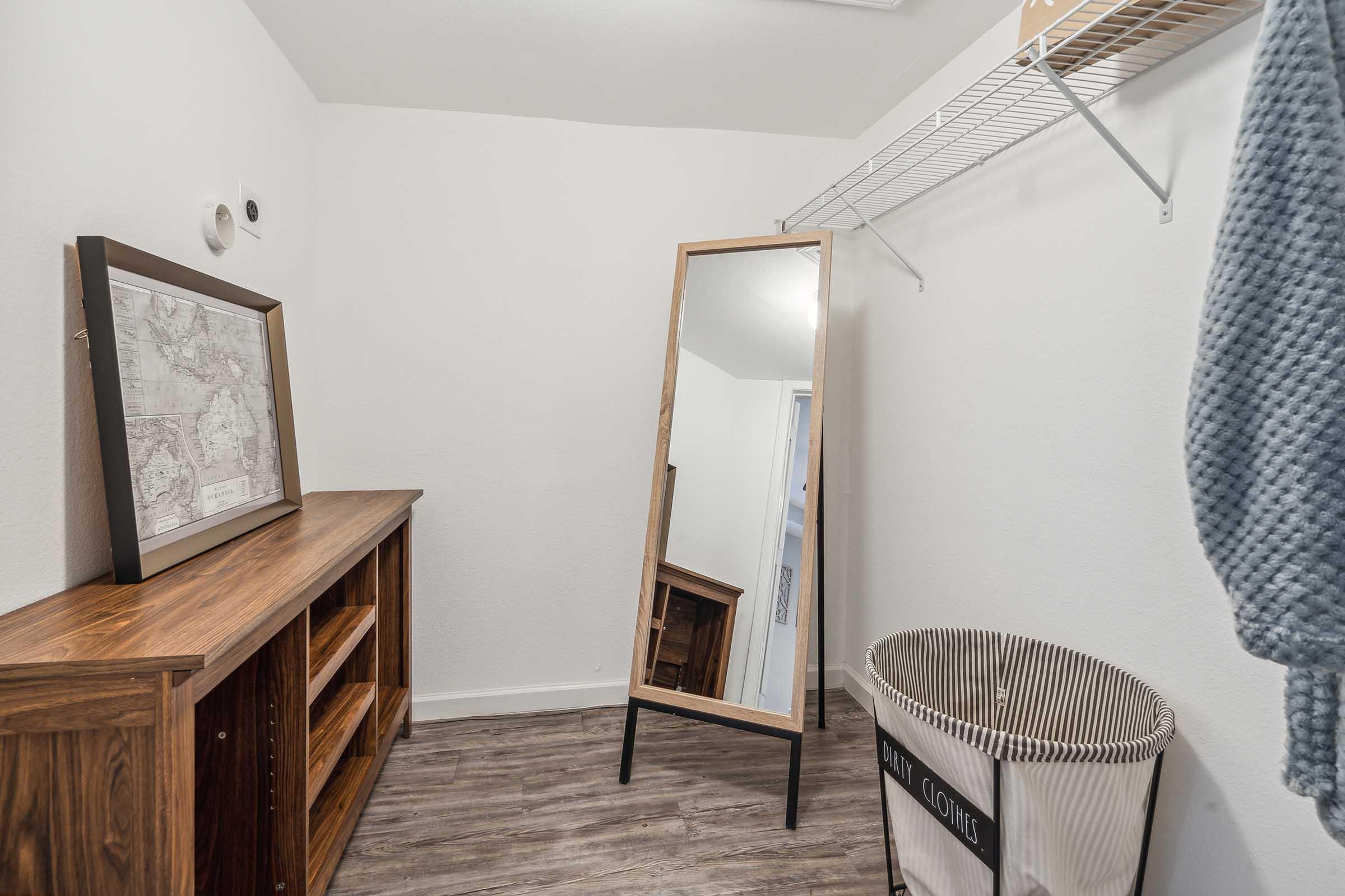 A small, organized room featuring a tall mirror, a wooden cabinet with shelves, a laundry basket, and a wall-mounted clothing rack. The walls are painted white, and the floor has a wood-like finish, creating a clean and simple aesthetic.