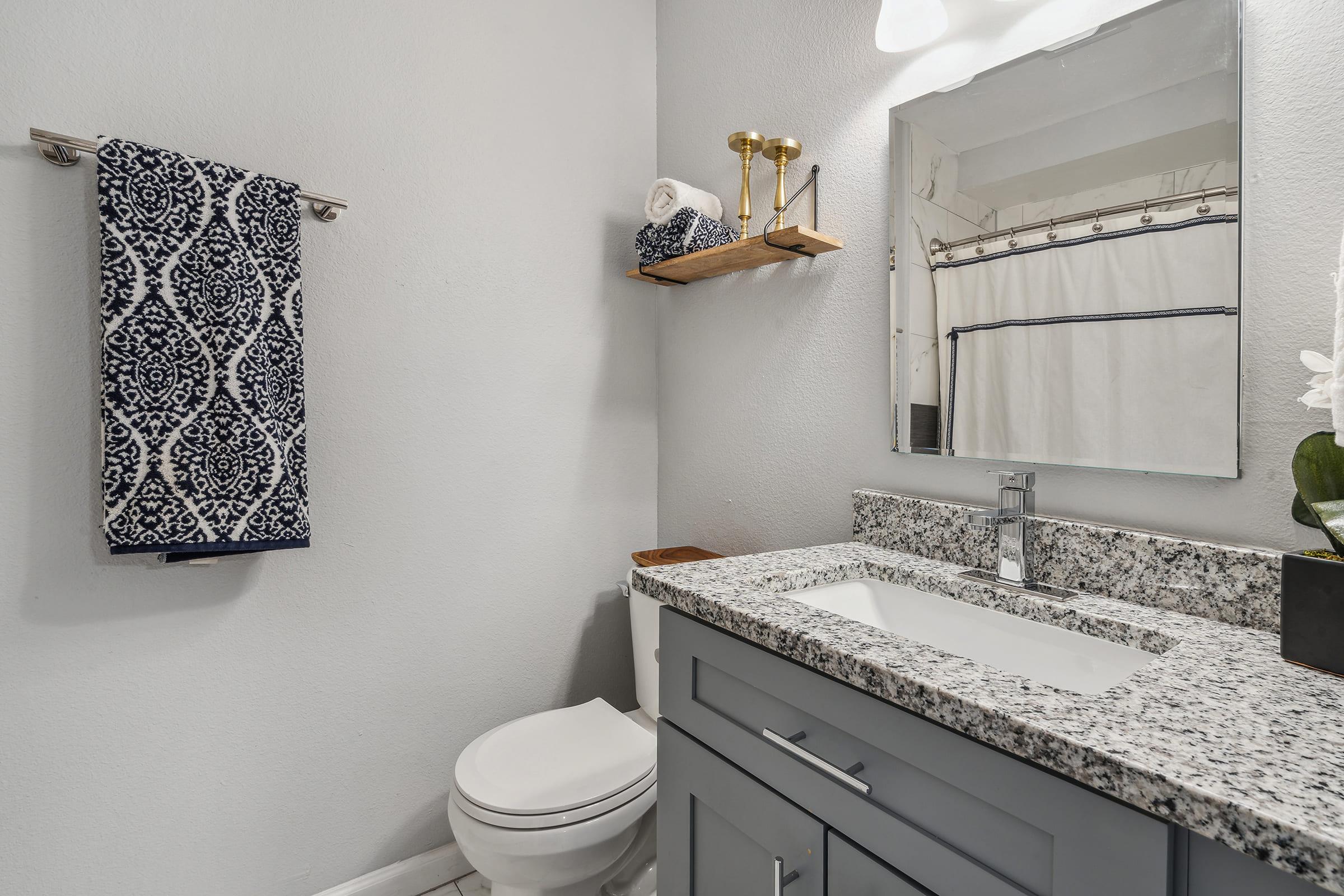 A modern bathroom featuring a granite countertop sink, a wall-mounted mirror, a white toilet, and a towel rack with a blue patterned towel. A small wooden shelf holds neatly stacked towels and a decorative plant. The shower curtain is neutral-toned, complementing the light grey walls.