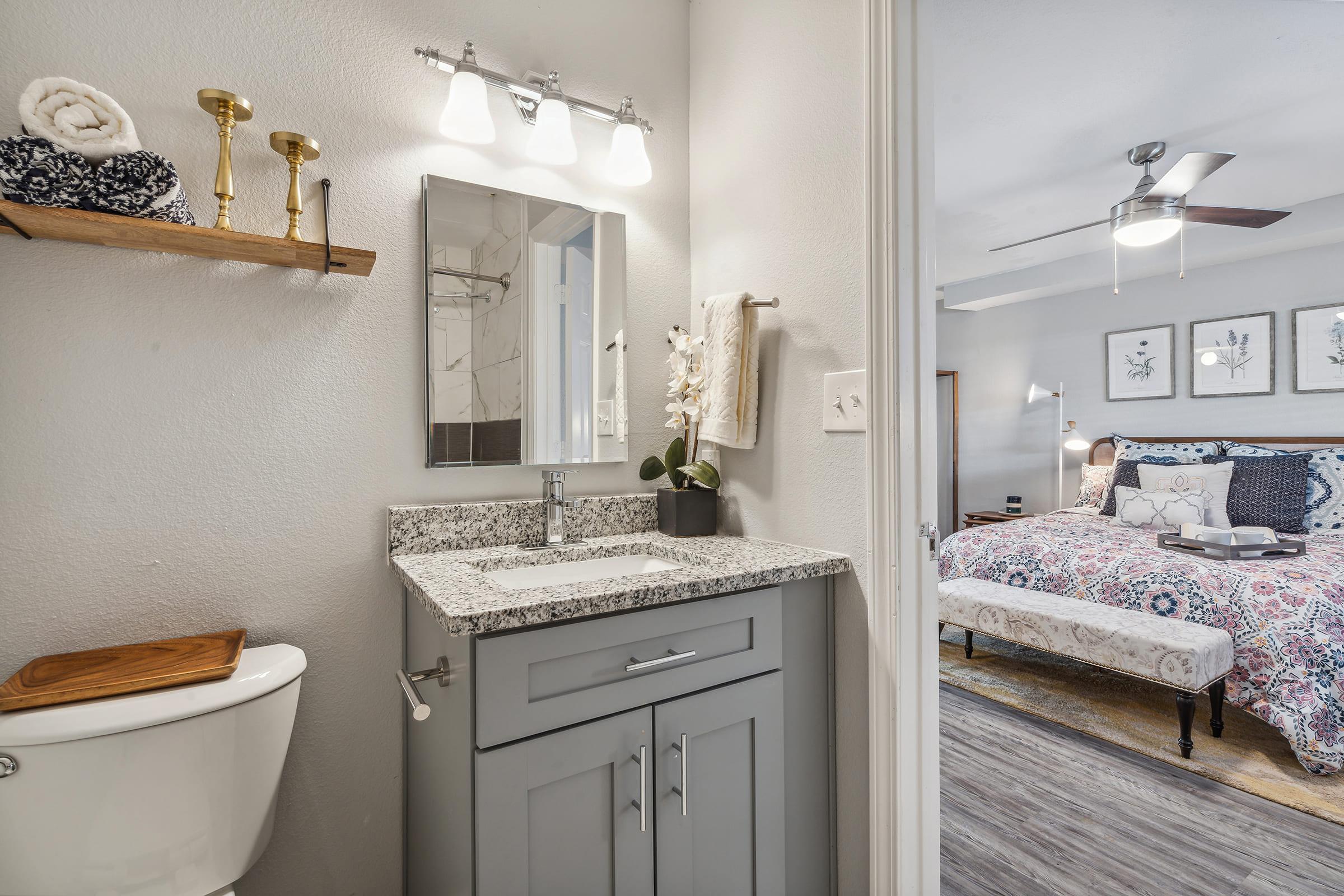 A modern bathroom featuring a gray vanity with a granite countertop, a mirror above, and a neatly arranged shelf. There is a white toilet and a view into a stylish bedroom with a ceiling fan and a patterned bedspread, adding a touch of elegance to the space.