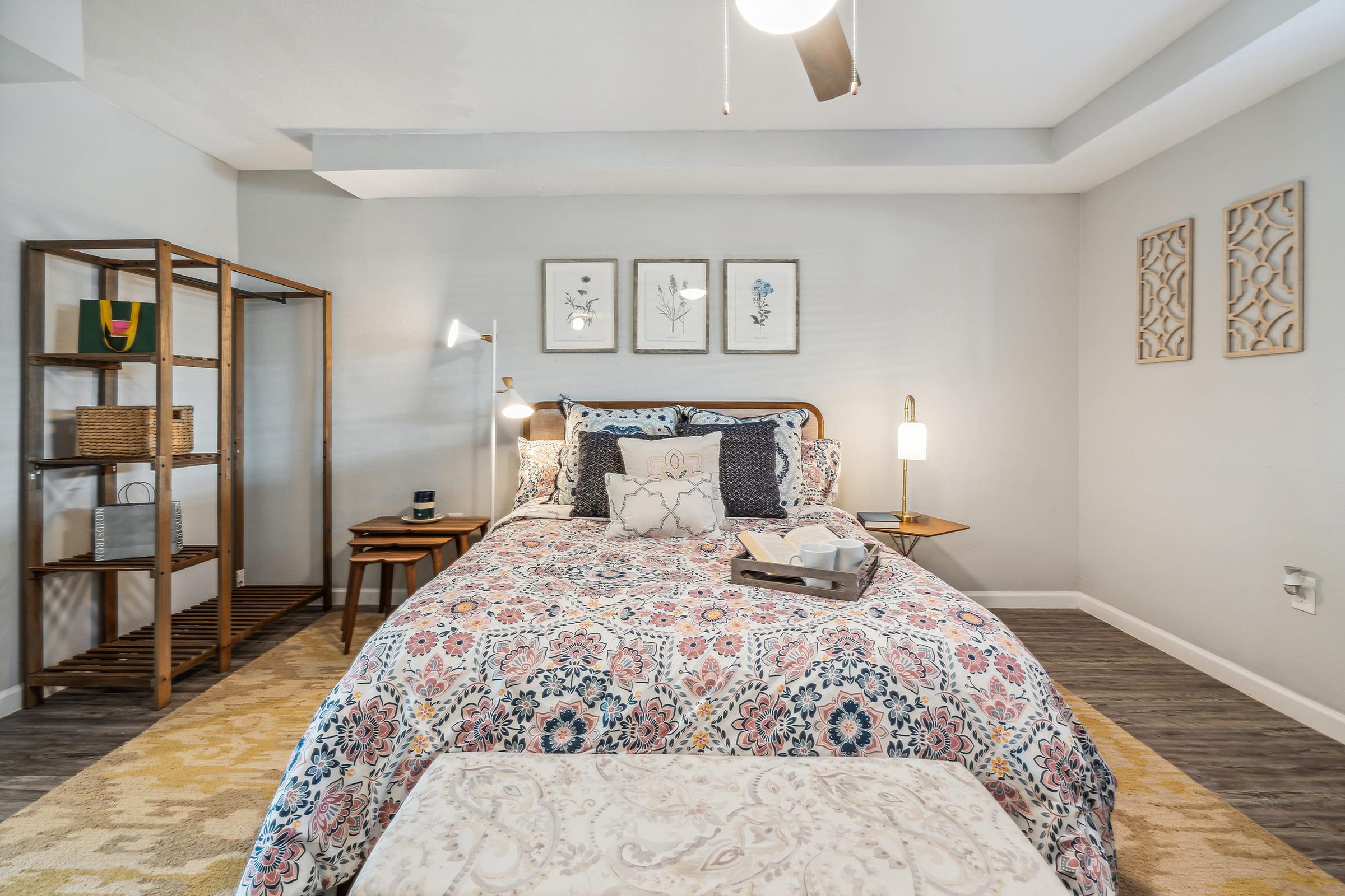 A cozy bedroom featuring a patterned bedspread on a wooden bed, complemented by decorative pillows. There are two lamps on bedside tables, a tray with items on the bed, and a wooden shelf with storage baskets. Wall art and subtle decor enhance the inviting atmosphere.