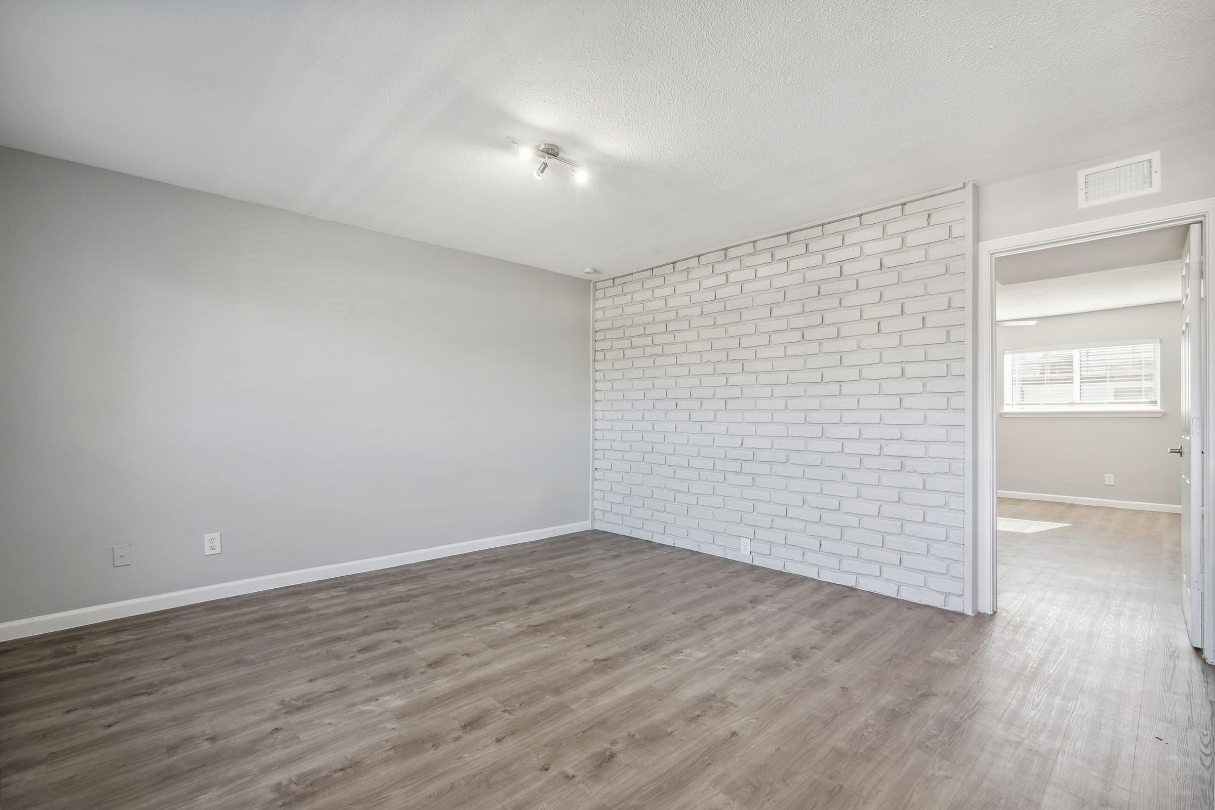 A spacious, empty room with light gray walls and a white brick accent wall. The flooring is light wood, and there are two windows allowing natural light in. A ceiling light fixture is present, and a door is visible on the right, leading to another room or area. The overall appearance is clean and minimalistic.