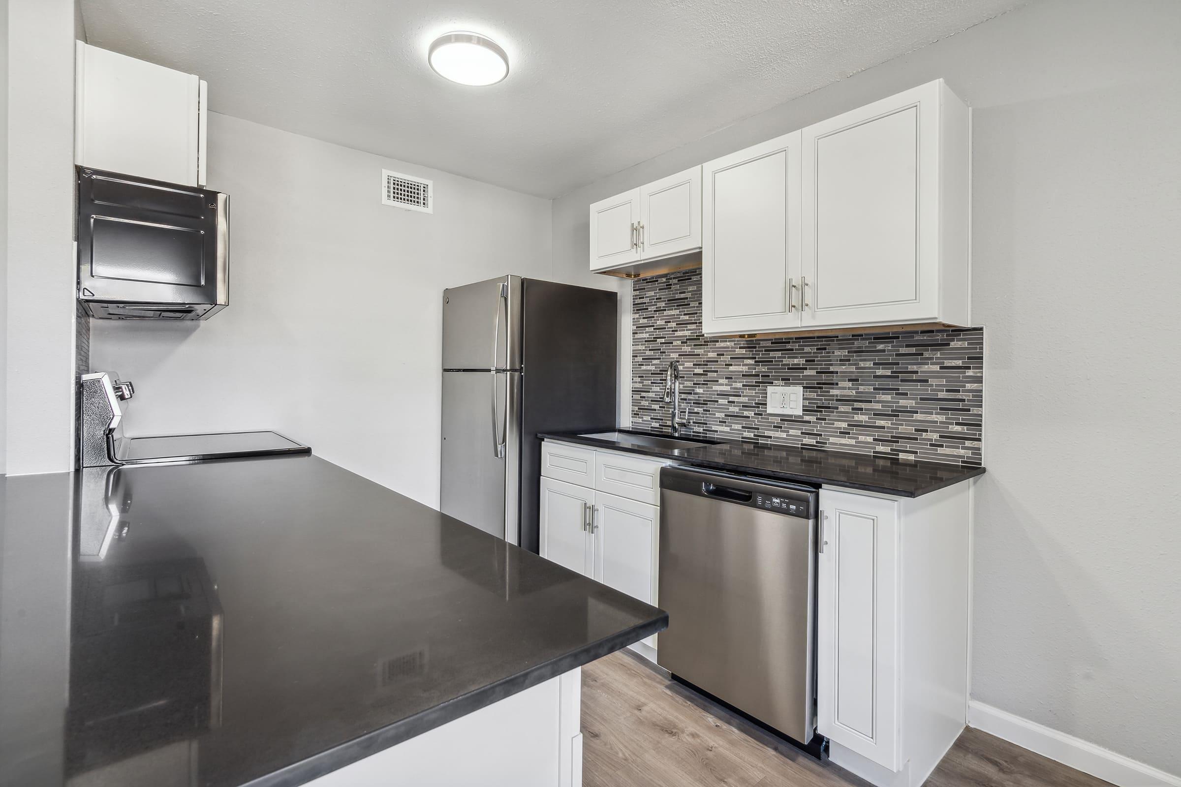 Modern kitchen featuring white cabinetry, stainless steel appliances including a refrigerator, dishwasher, and a stove. The countertop is dark and smooth, complementing the light gray walls. The backsplash consists of small, colorful tiles that add texture and style to the space.