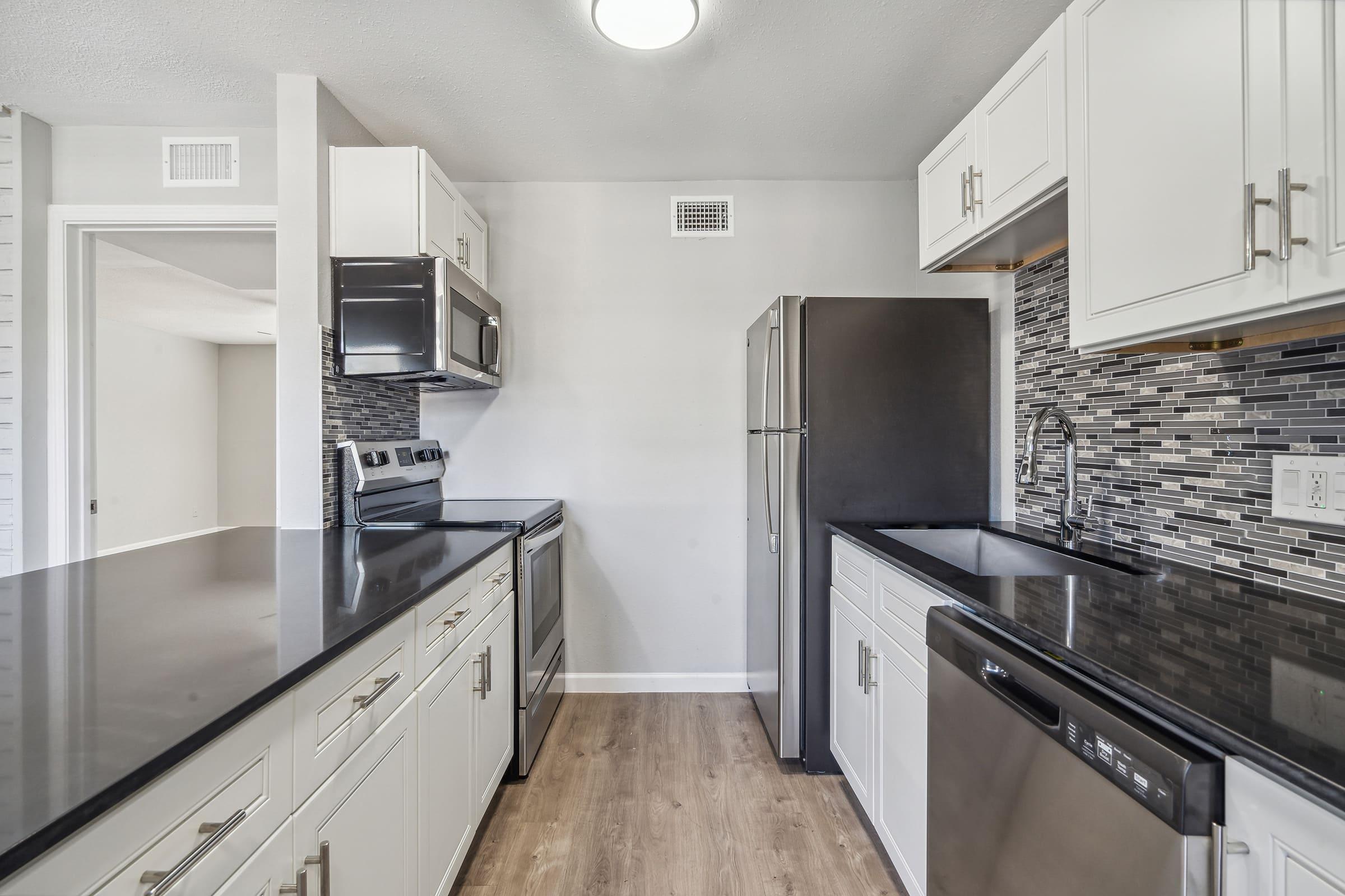Modern kitchen with white cabinets, dark countertops, and stainless steel appliances, including an oven, microwave, refrigerator, and dishwasher. A tiled backsplash adds a stylish touch. The design features an open layout with light-colored walls and wooden flooring.