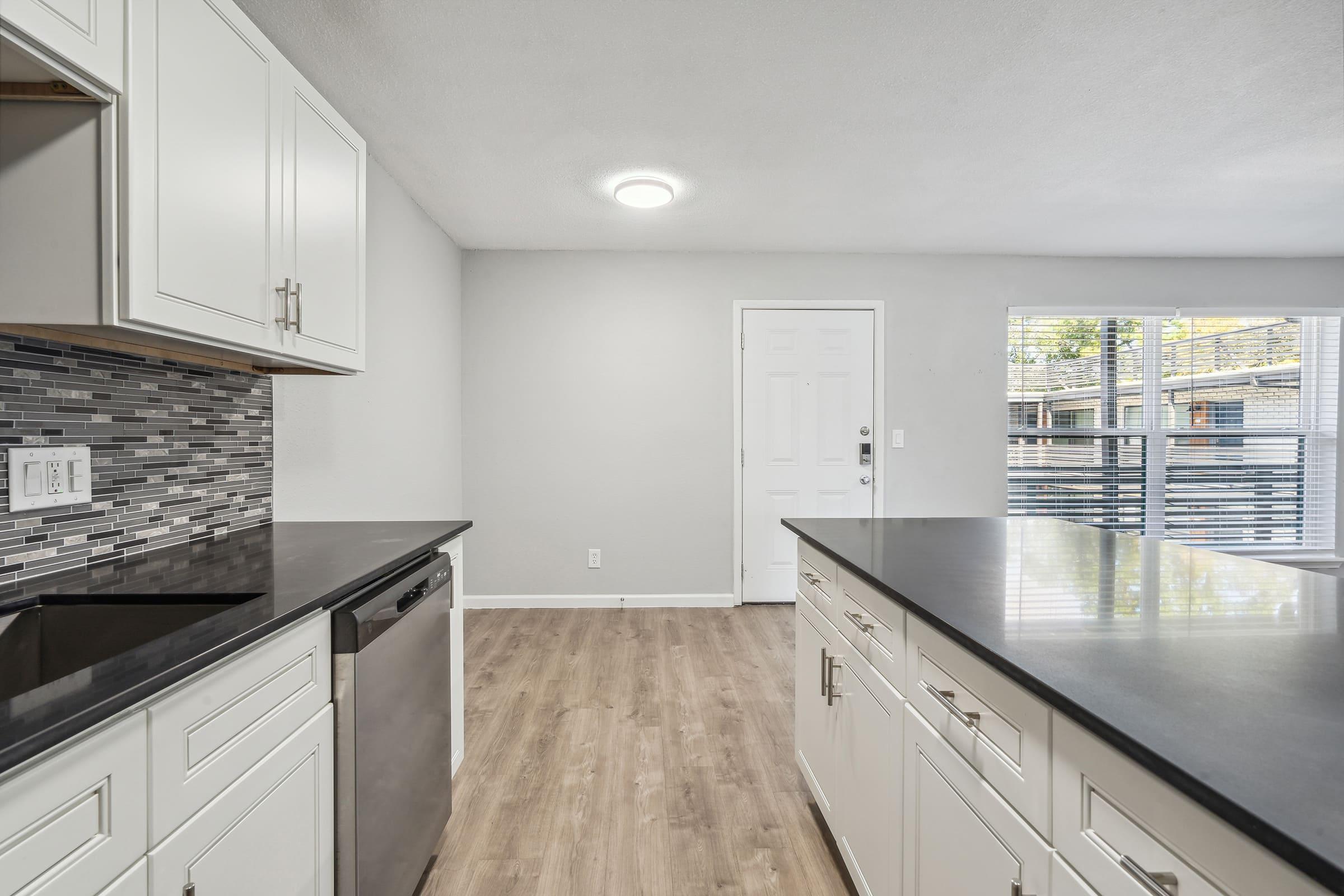 Modern kitchen interior showing a view of white cabinetry, a stainless steel dishwasher, and black countertops. Features include a mosaic tile backsplash, gray walls, a front door, and a window with outside view. The space has a warm, inviting atmosphere with natural light.