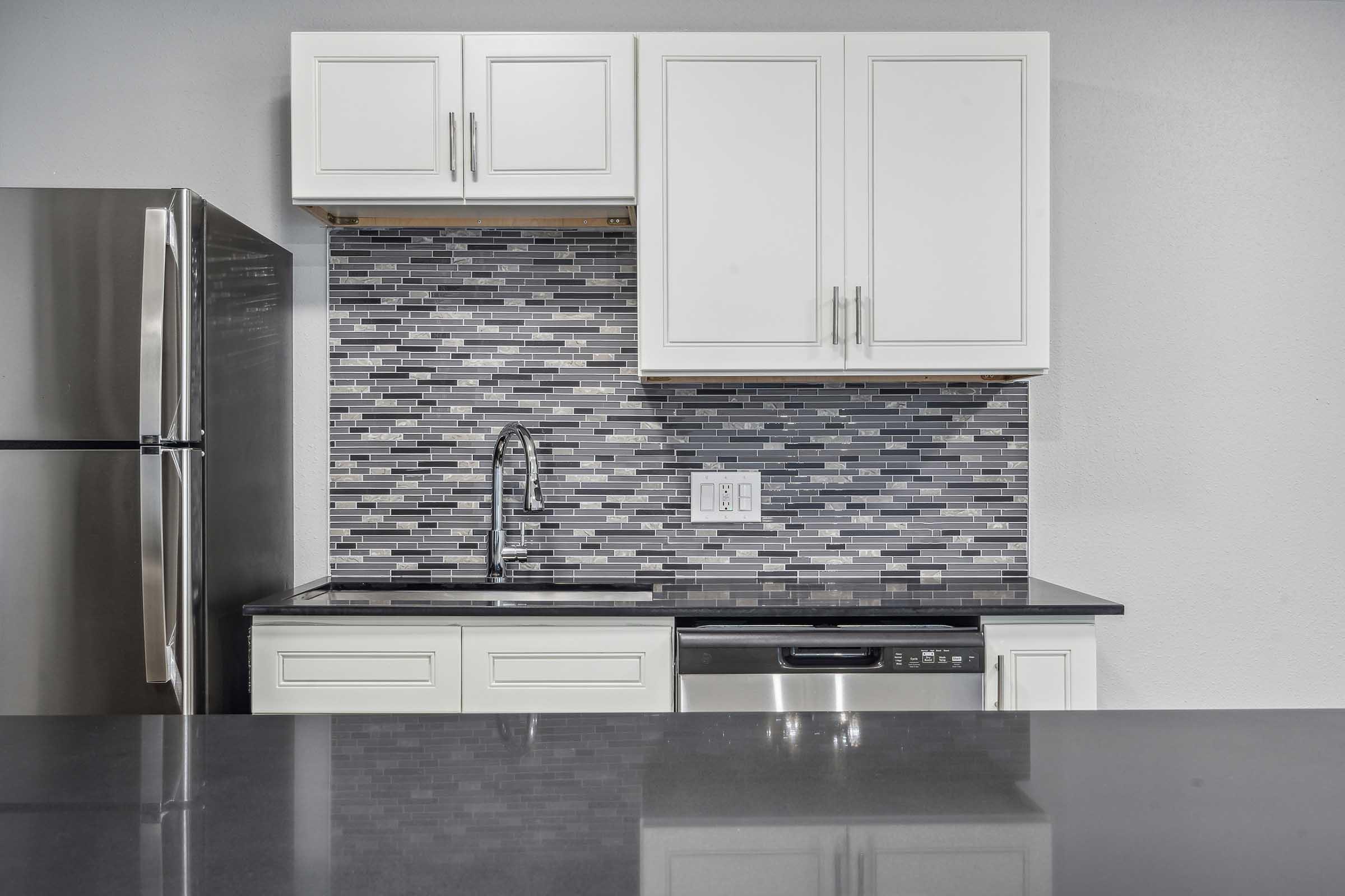 Modern kitchen featuring sleek white cabinets, a stainless steel refrigerator, and a black countertop. The backsplash is composed of grey and black tiles, with a stainless steel sink and dishwasher integrated into the cabinetry. The overall design conveys a clean and contemporary aesthetic.