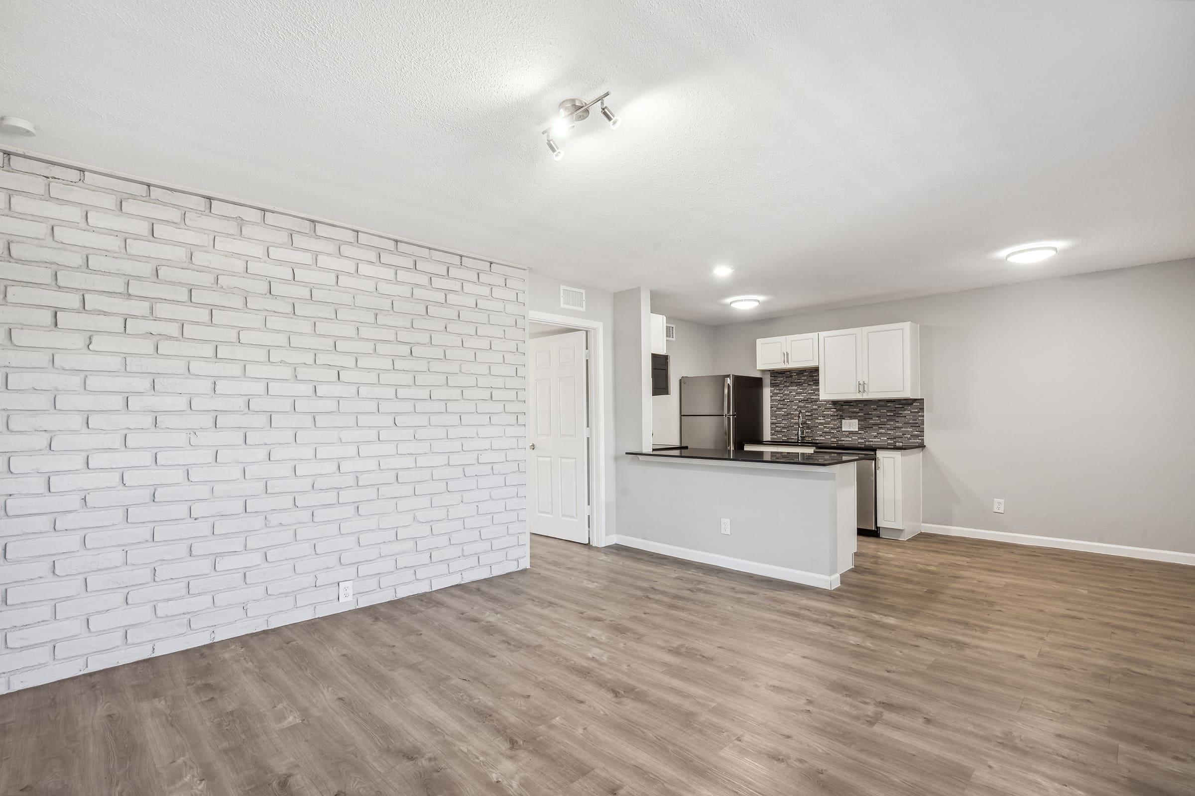 Spacious interior of a modern apartment featuring a light-colored brick accent wall, laminate flooring, and an open kitchen with white cabinetry and stainless steel appliances. The layout is bright and inviting, with natural light enhancing the space.