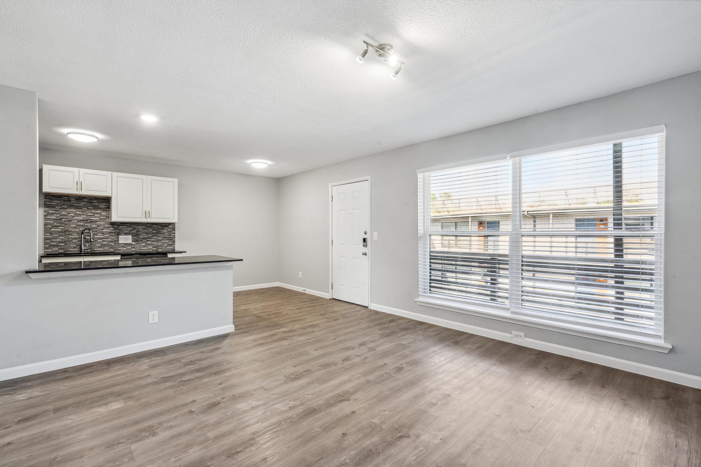 A spacious, modern living area featuring light gray walls, new laminate flooring, and an open kitchen with white cabinets and a dark countertop. Large windows with white blinds provide natural light, and a front door leads outside. The layout creates a bright and airy atmosphere.