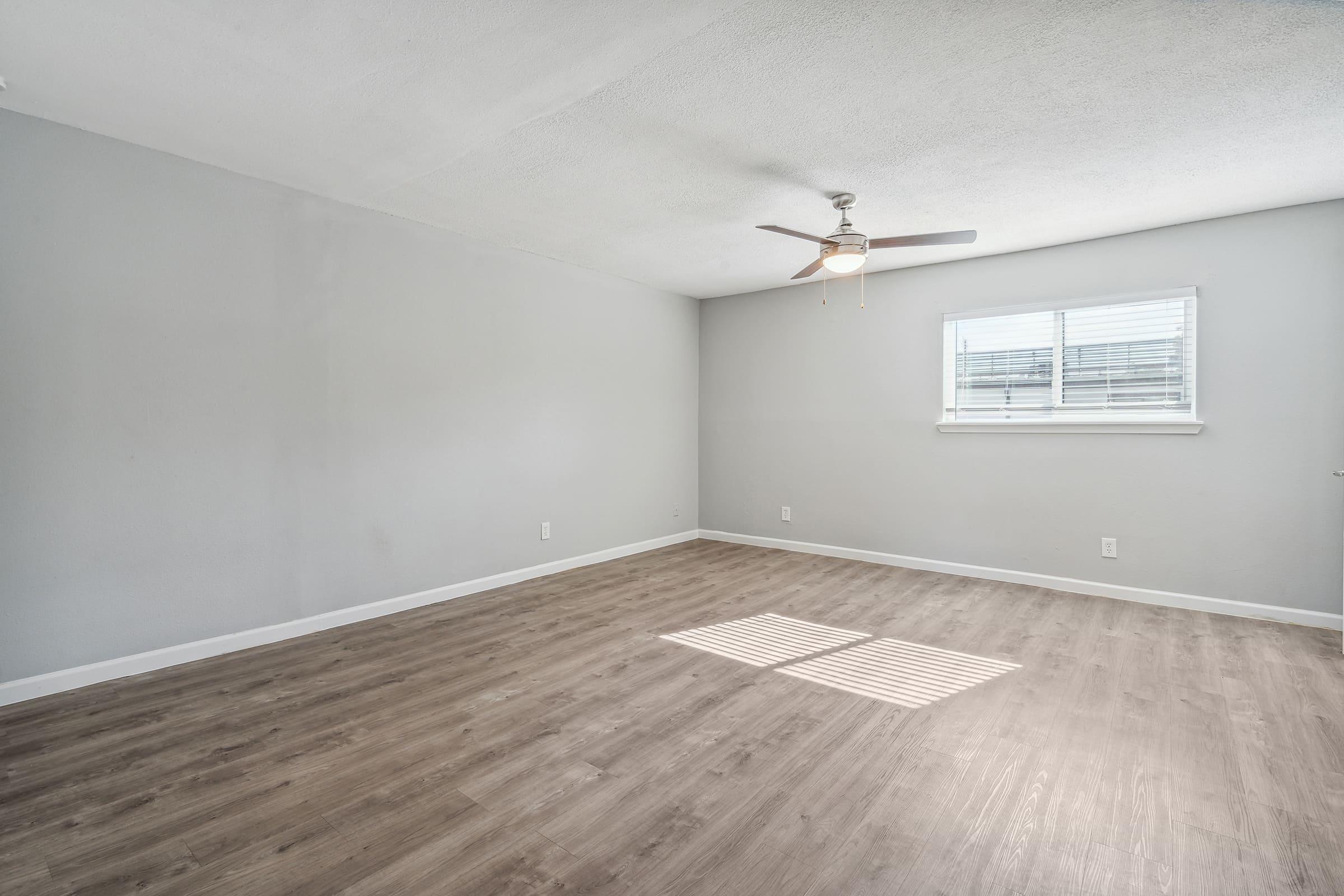 A spacious, empty room with light gray walls and light wooden flooring. A ceiling fan is installed, and a window with white blinds allows natural light to illuminate the space. The room appears clean and ready for furnishings.
