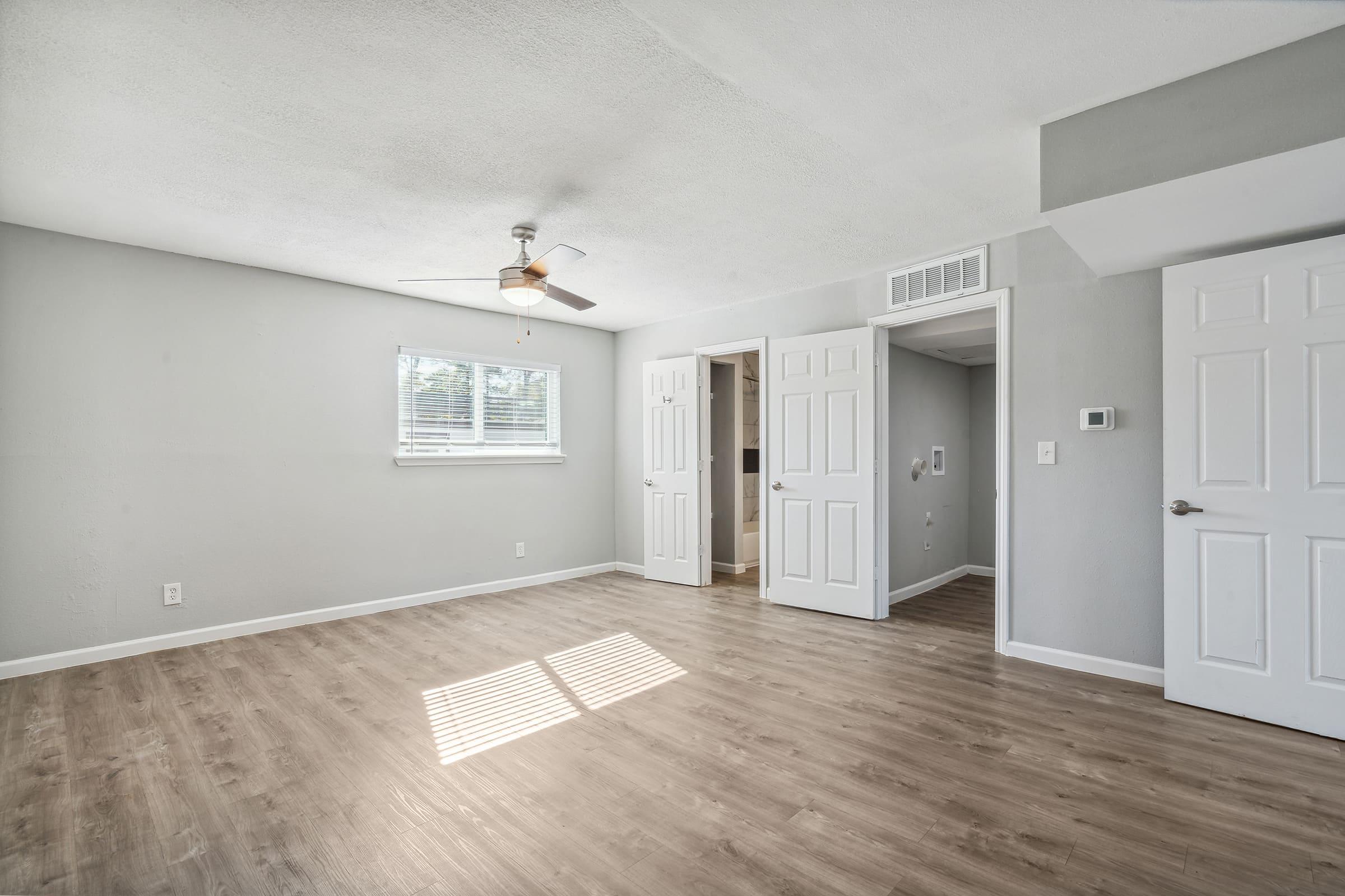 A bright, spacious room featuring light gray walls and wood-like flooring. There is a ceiling fan installed, and natural light streams in through a window. Two doors are visible: one leading to a closet and another to a separate area, creating an airy, inviting atmosphere.