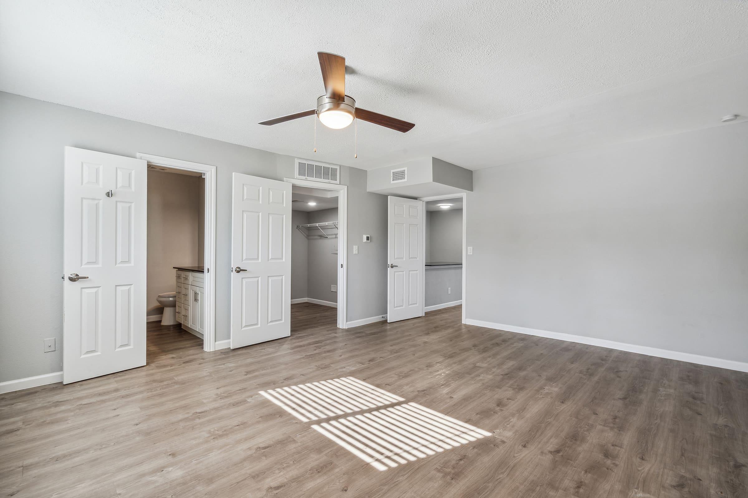 Spacious, well-lit room showcasing light wood flooring. The area features two open doors leading to separate spaces. One door opens to a bathroom, while the other leads to a closet. A ceiling fan adds to the airy atmosphere, and natural light is visible through window shadows on the floor.