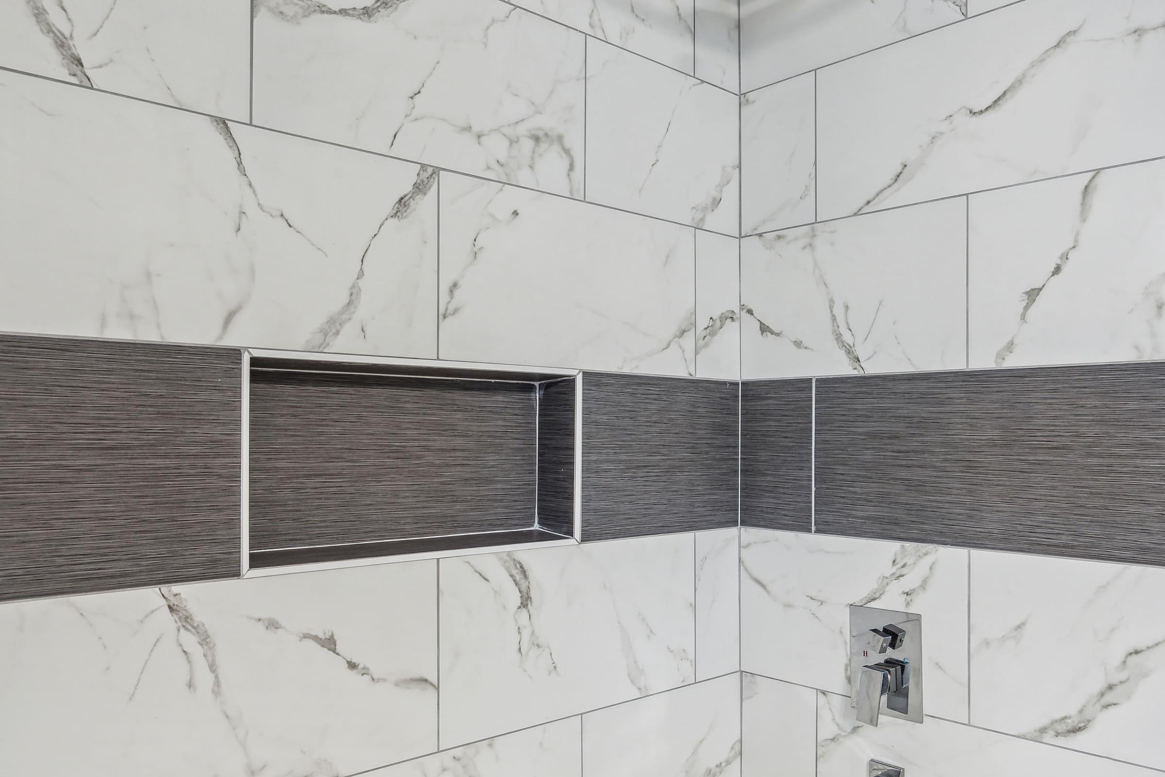 A modern shower wall featuring a combination of white marble tiles and textured gray tiles. The design includes a recessed shelf for storage, and a sleek faucet installed below. The overall aesthetic is clean and contemporary, emphasizing the contrast between the materials.