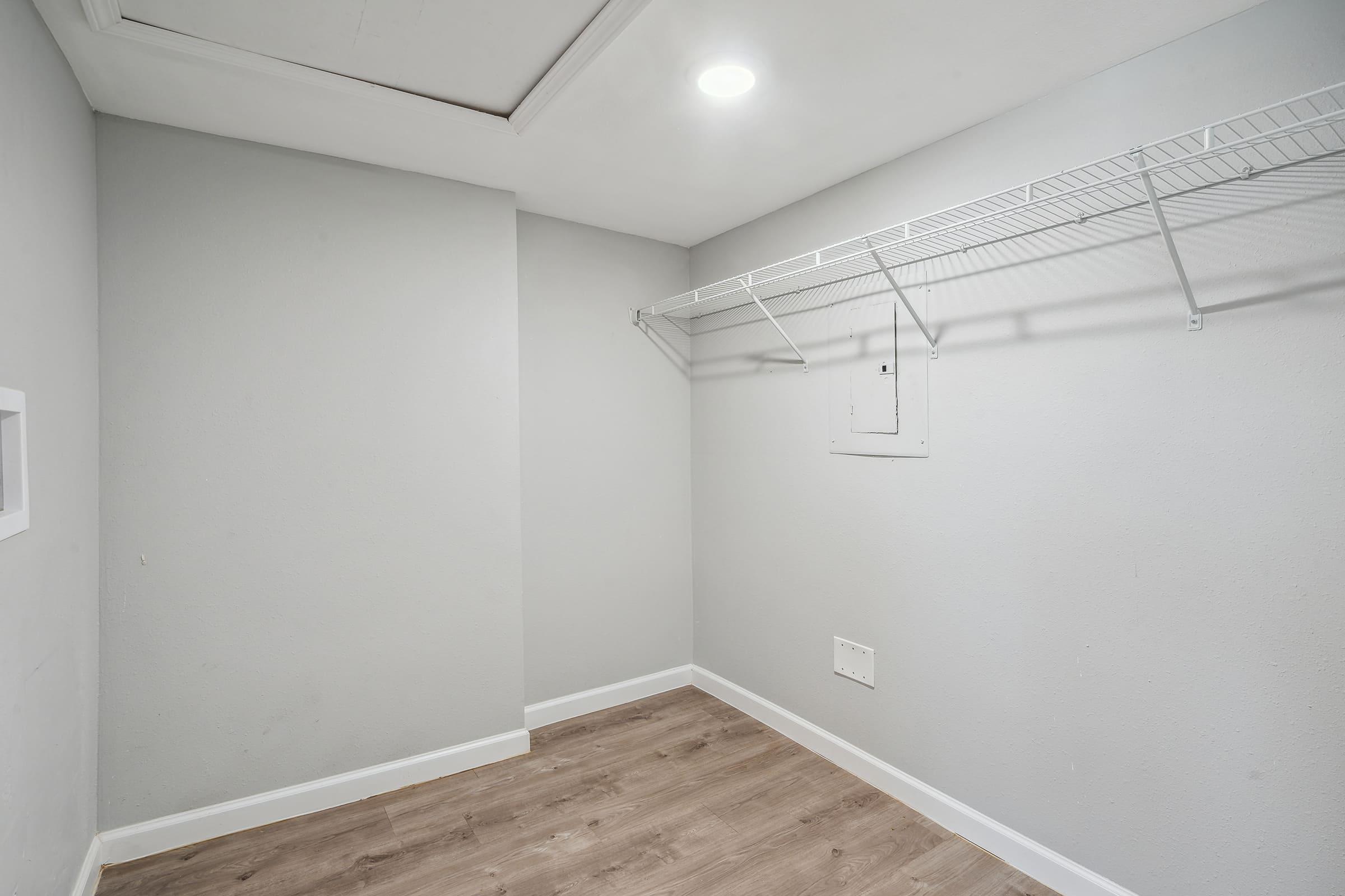 Empty closet with light gray walls and a light-colored wooden floor. There is a wire shelf along one wall and a small electrical panel on another wall. The ceiling features a subtle overhead light, creating a bright, spacious feel despite the minimal furnishings.